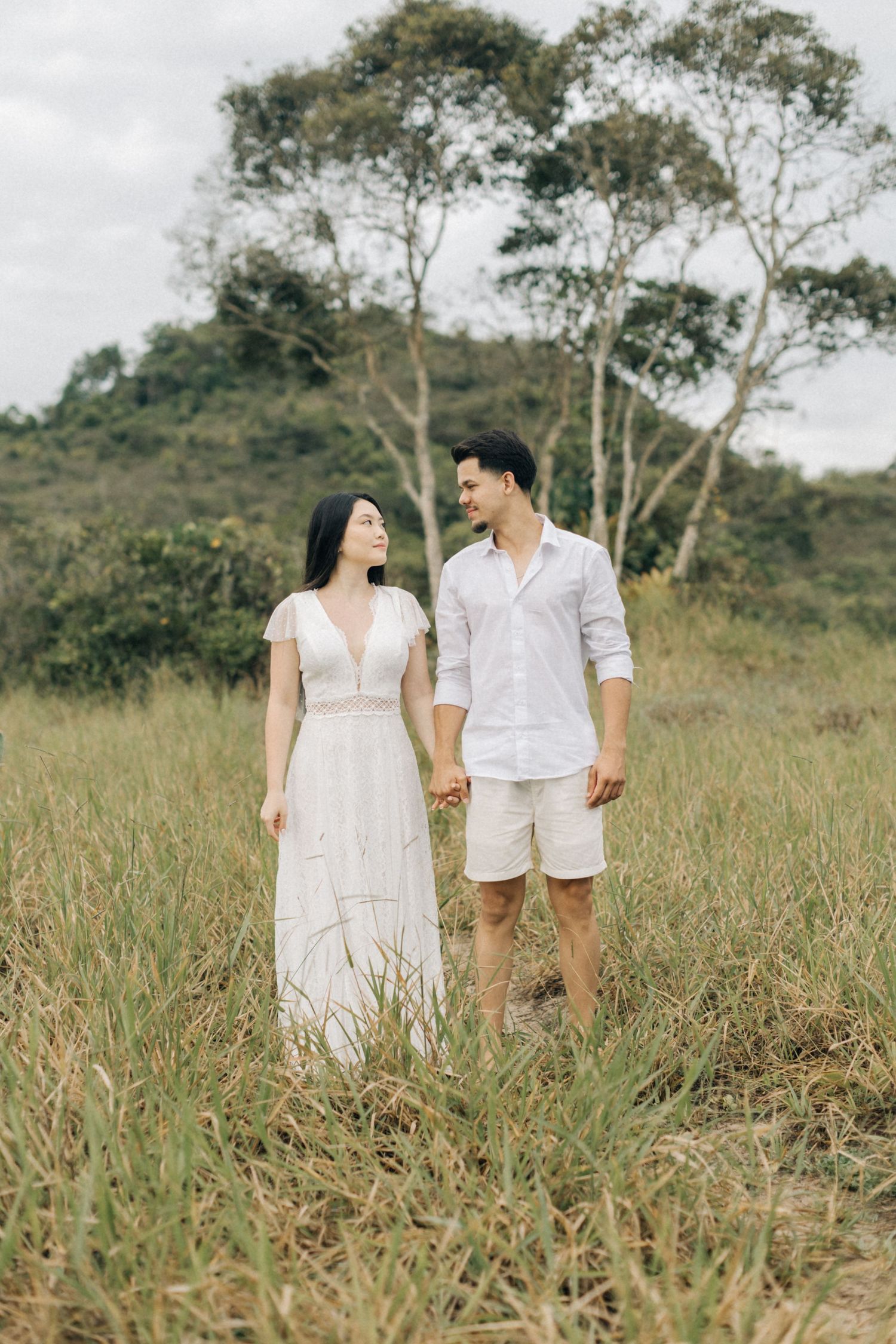 A couple stands together in a meadow wearing matching white outfits against a hillside backdrop.