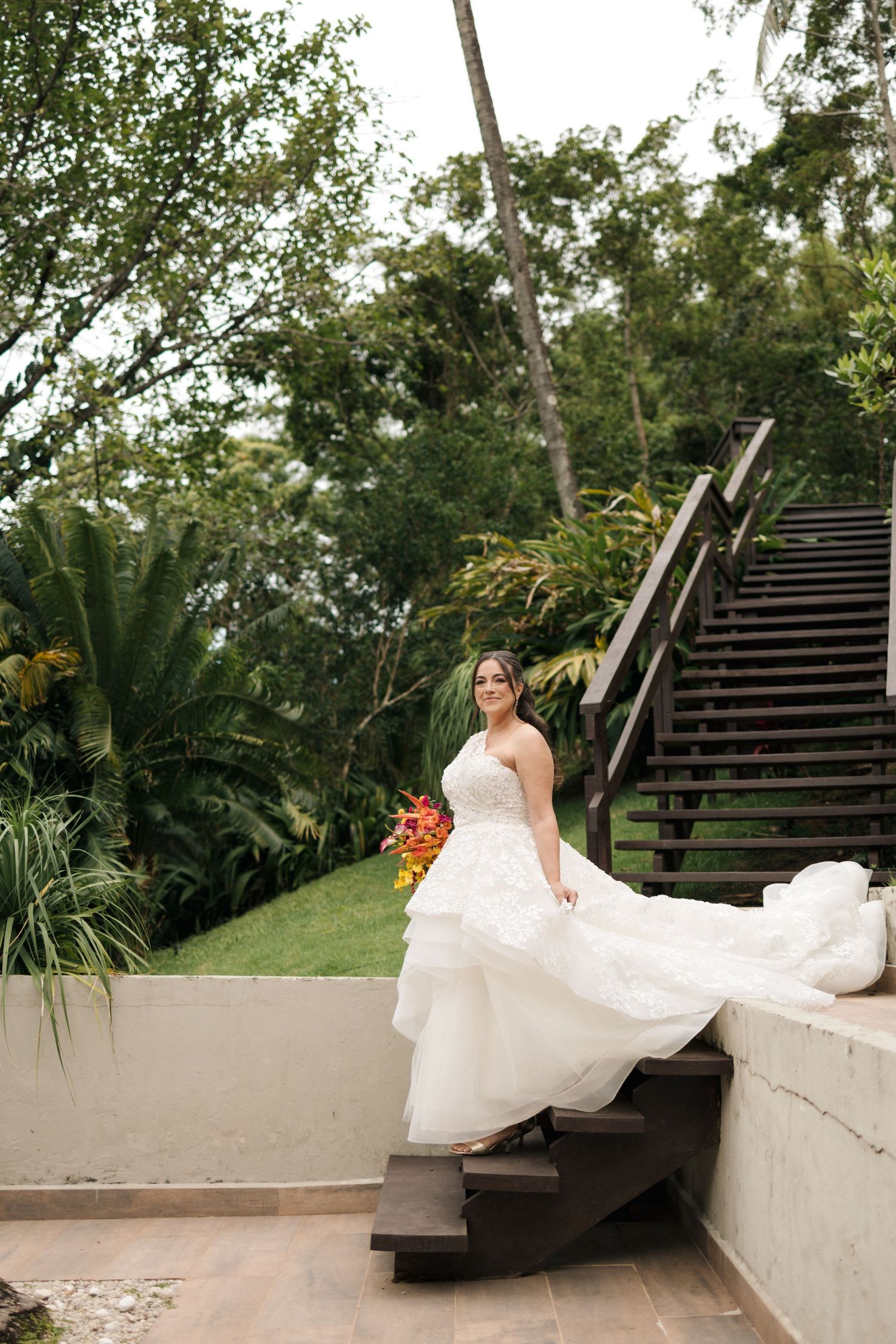 A bride in a flowing white dress poses on outdoor steps surrounded by tropical greenery and palm trees.