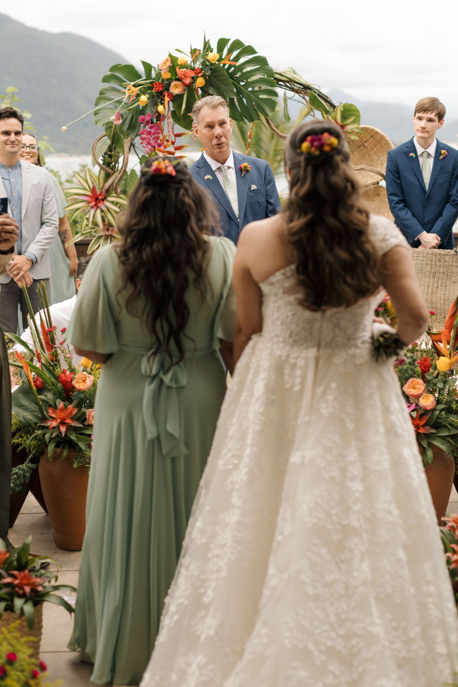 Wedding party stands against tropical backdrop with green monstera leaves and orange floral arrangements.