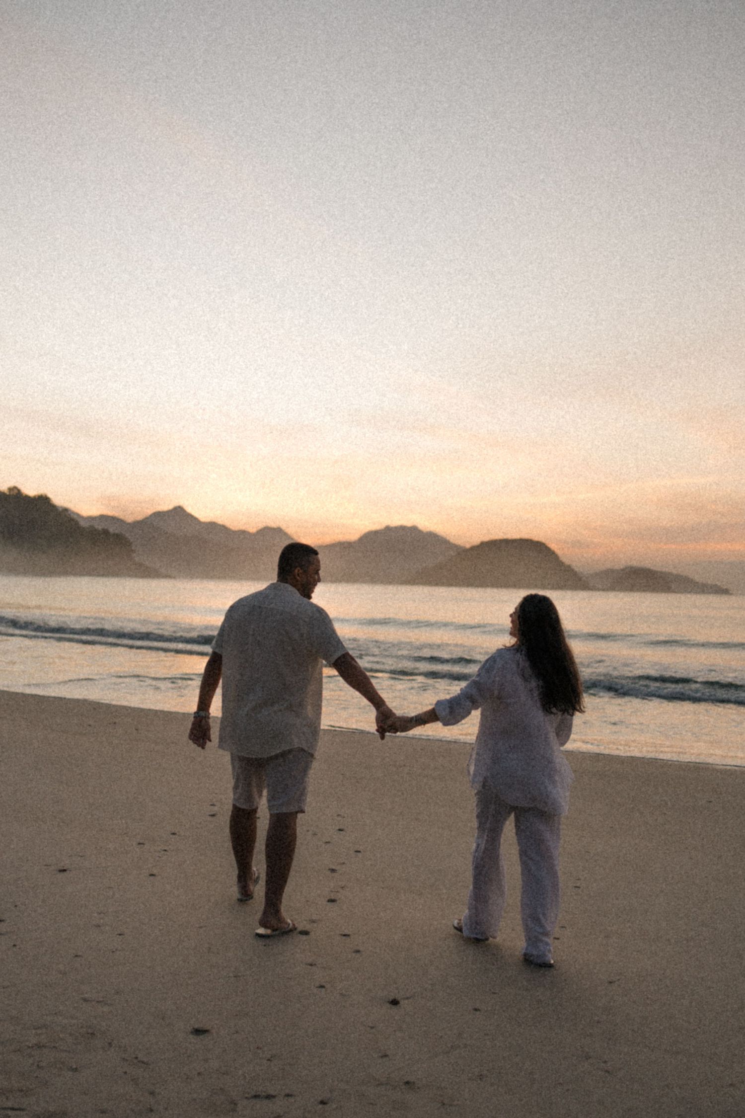Couple holding hands while walking on an empty beach during sunset with mountains in background.