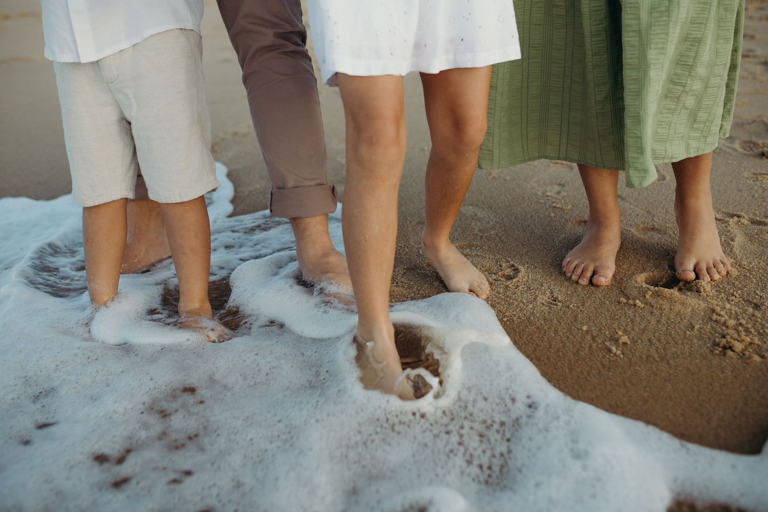 Feet and legs of people standing in beach sand with waves washing up on shore.