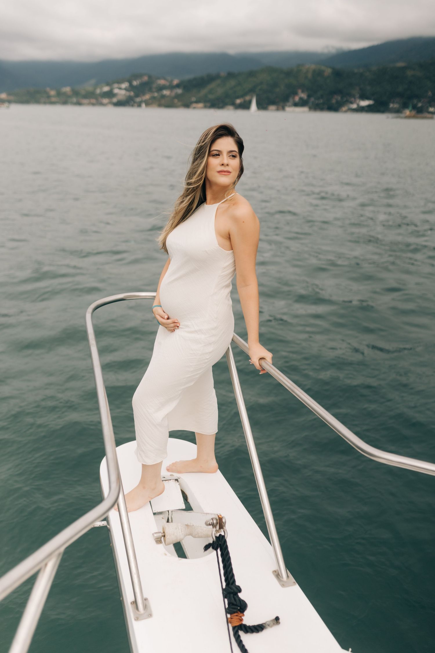 A woman in white dress poses gracefully on the edge of a boat with mountains in background.