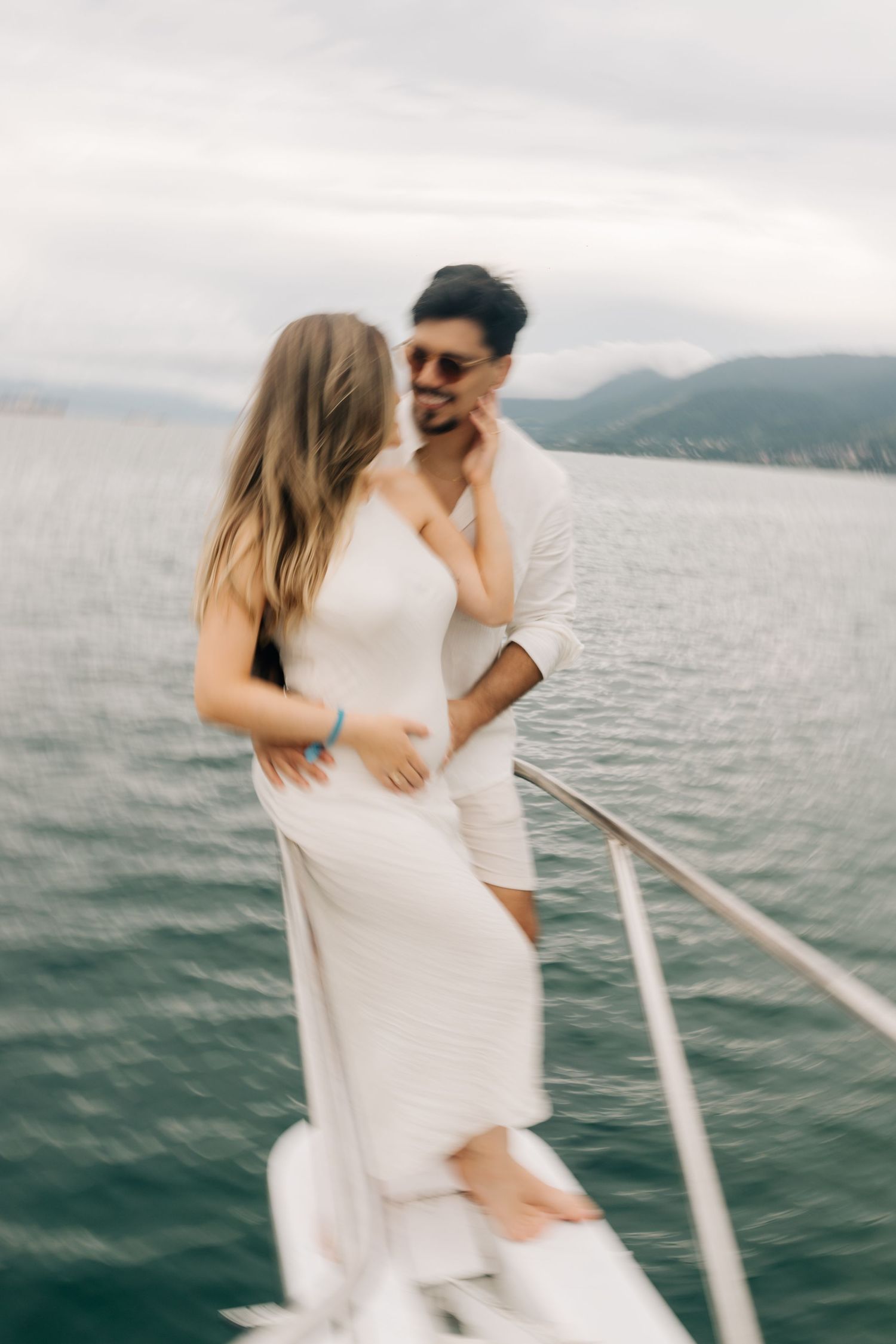 Couple in white sharing an intimate moment on a boat deck with mountains in background.