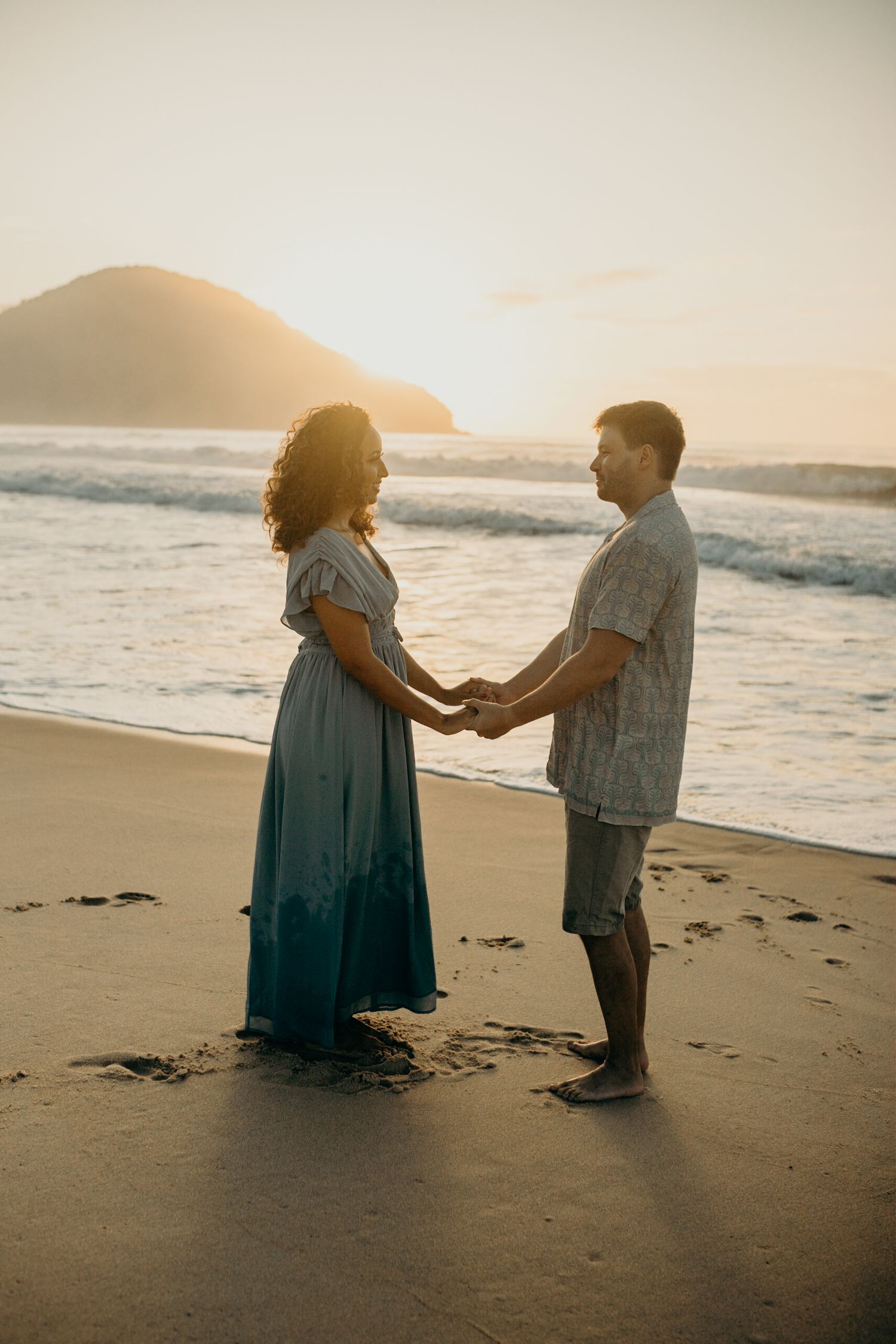 Couple holding hands while standing on beach at sunset with mountain backdrop.