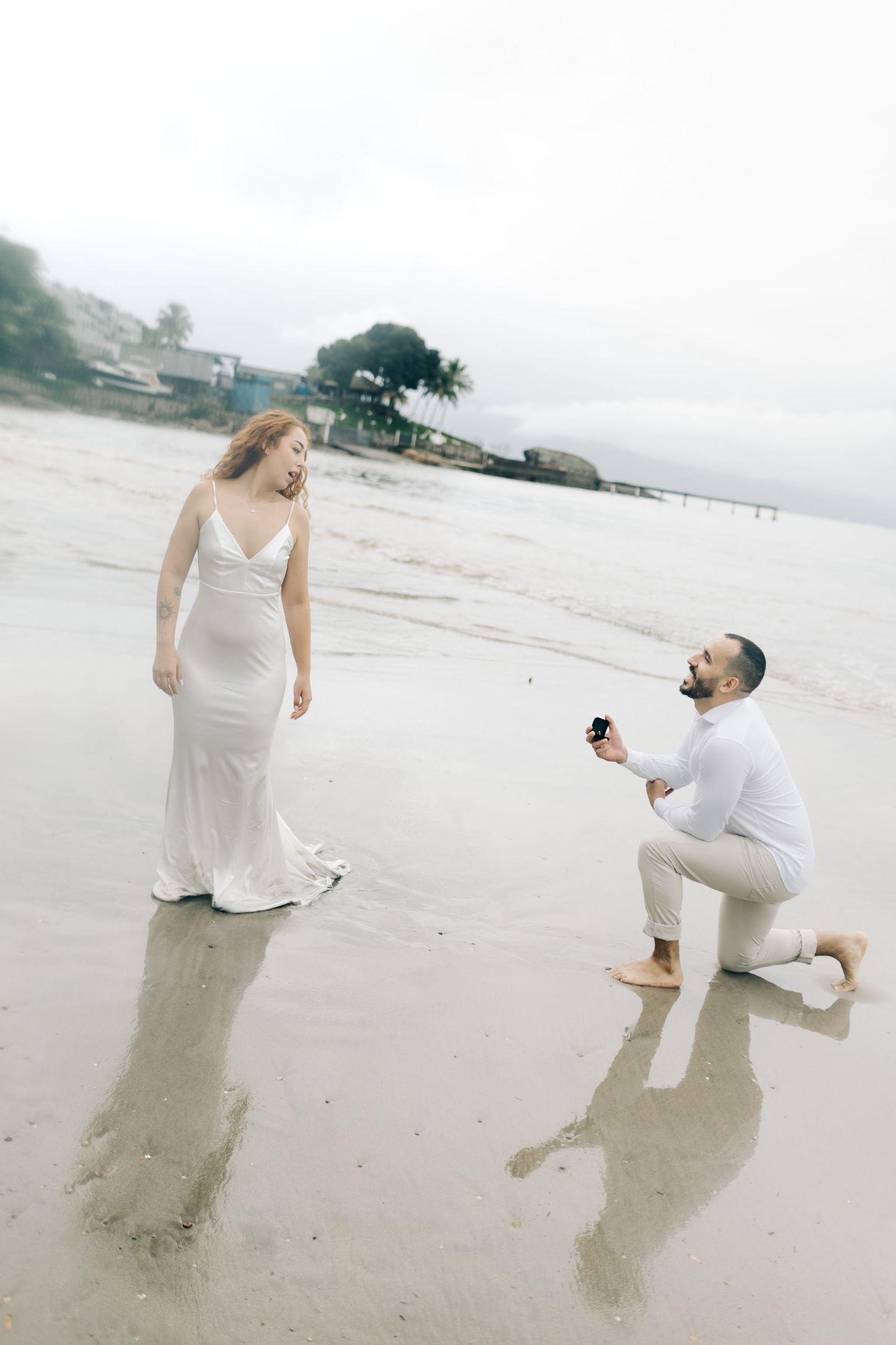 Romantic beach proposal with person in white dress and other kneeling with ring.