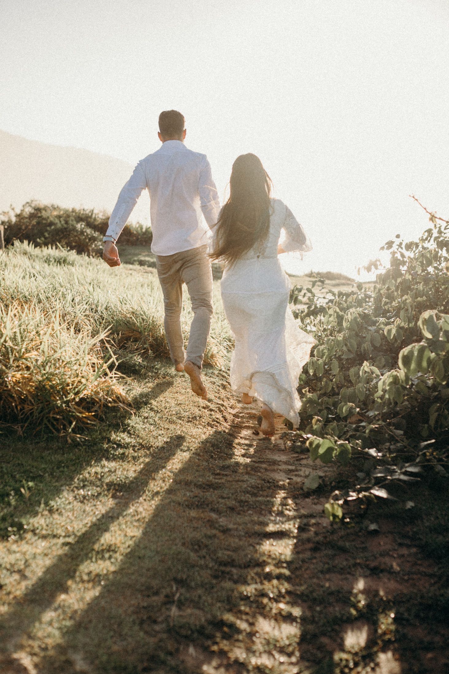 Couple walking hand in hand on sunlit hill path in wedding attire.