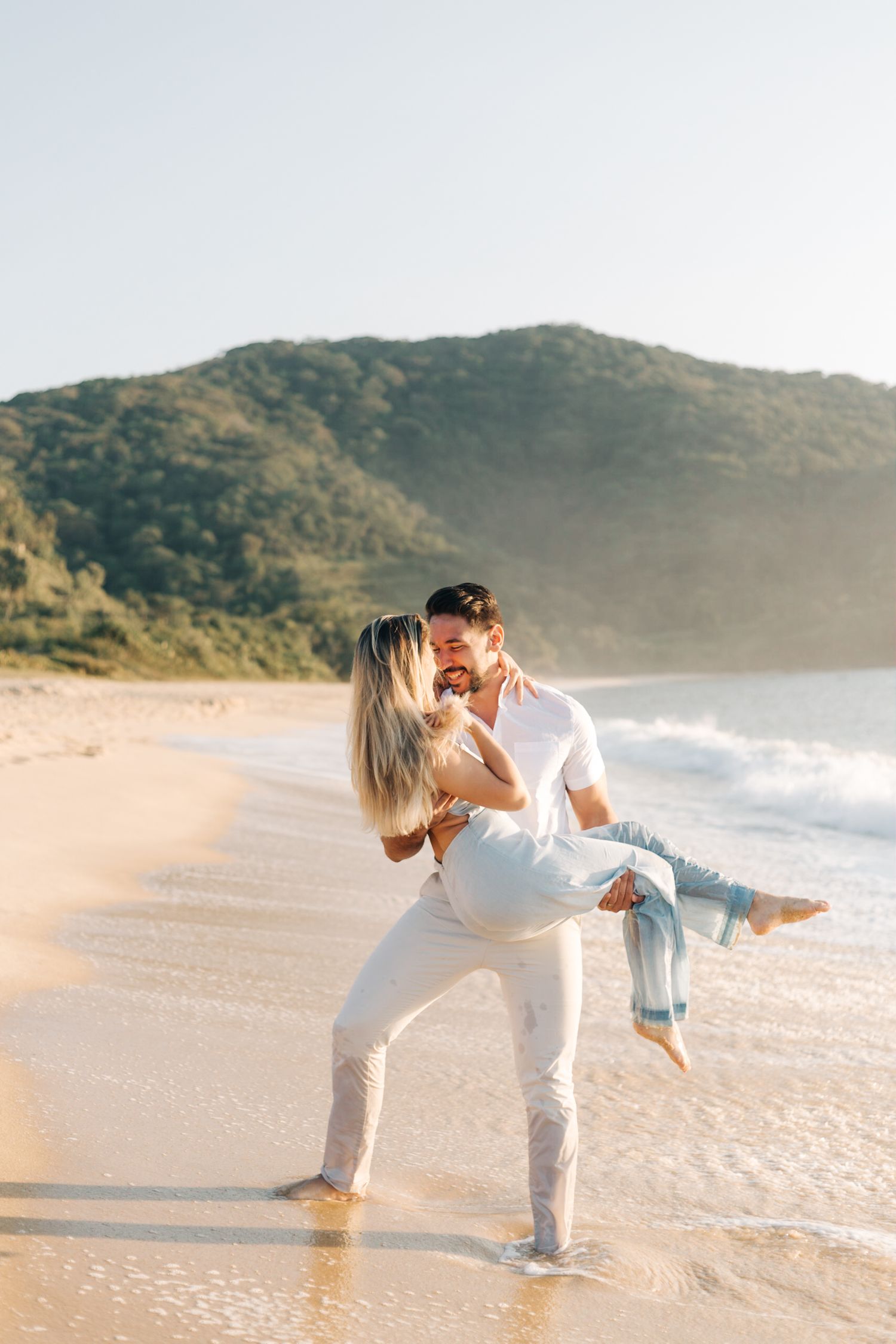 Couple dancing and laughing together on sunlit beach with mountain backdrop.