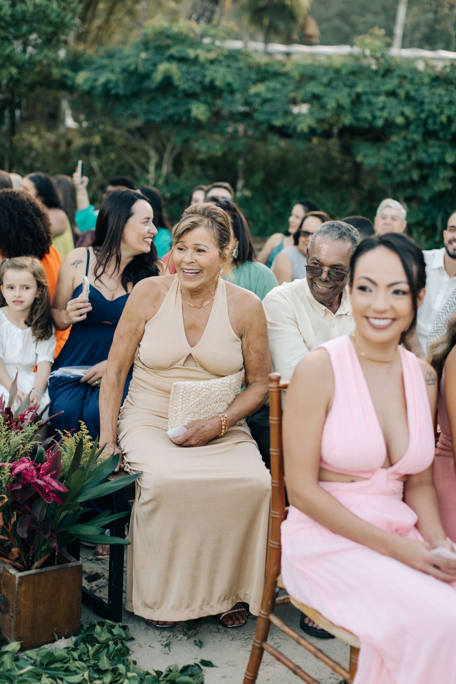 Wedding guests in pastel dresses seated outdoors among tropical flowers.