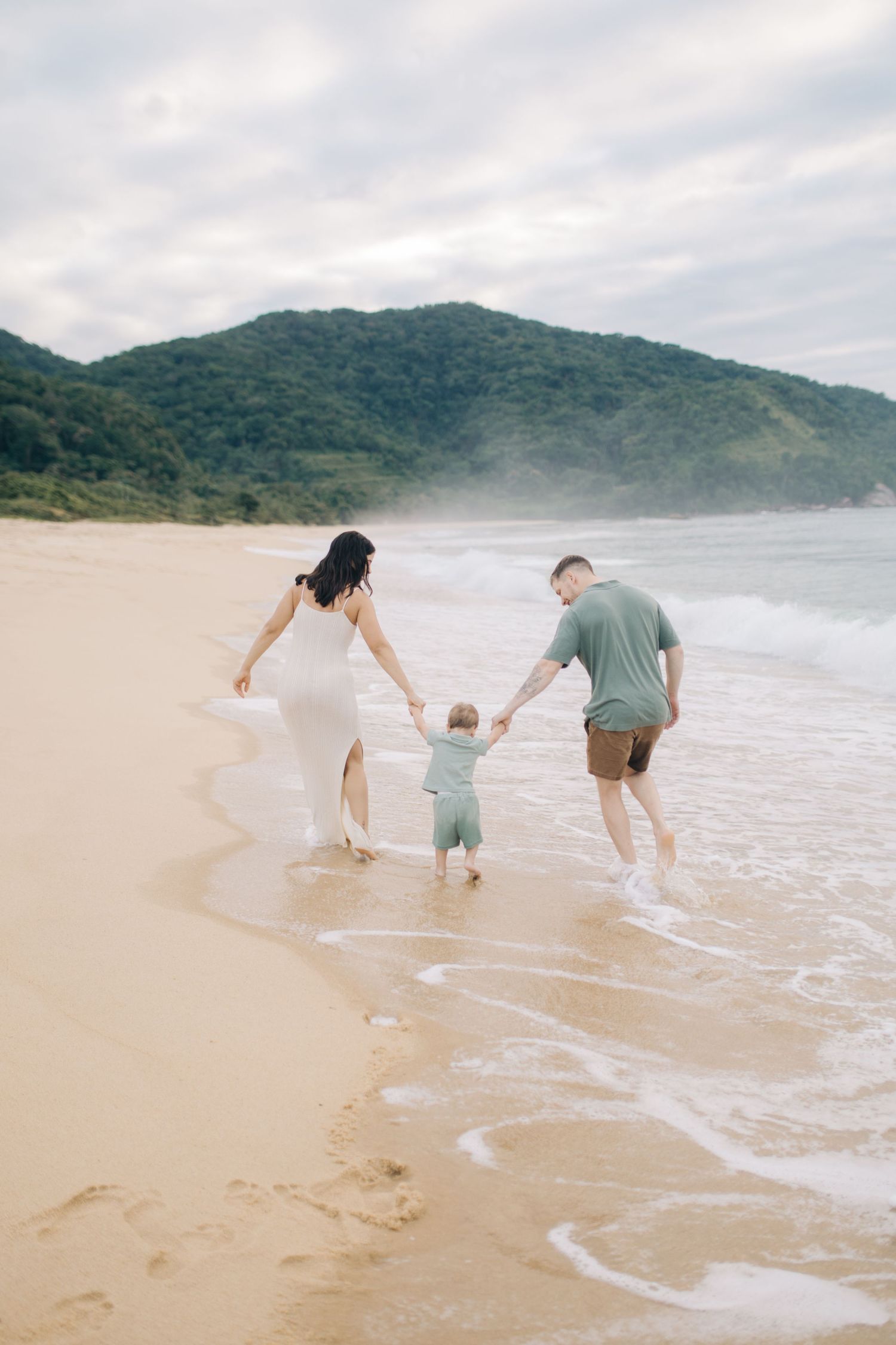 A family plays together at the beach's edge, chasing waves on a cloudy day.