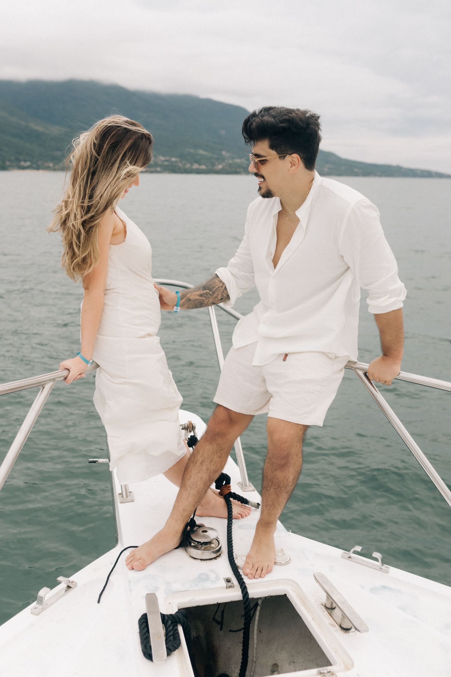 A couple in all white outfits share a playful moment on the bow of a boat.