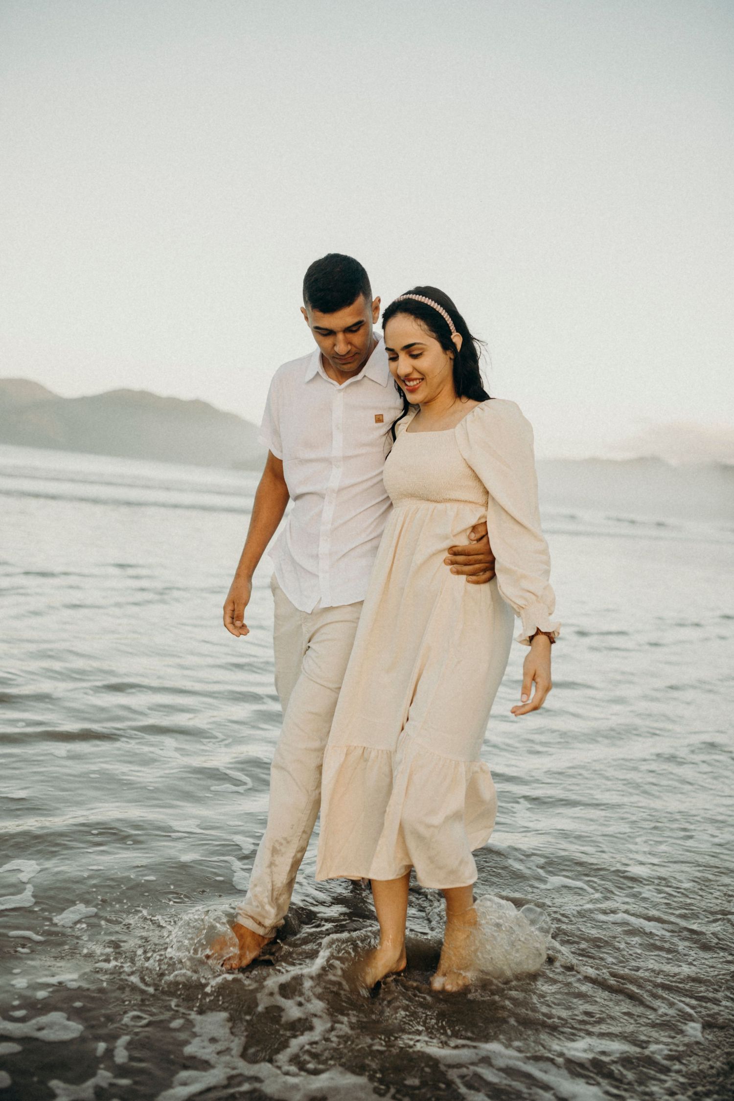 A couple in light colored outfits splashes playfully in the ocean waves at the shore.