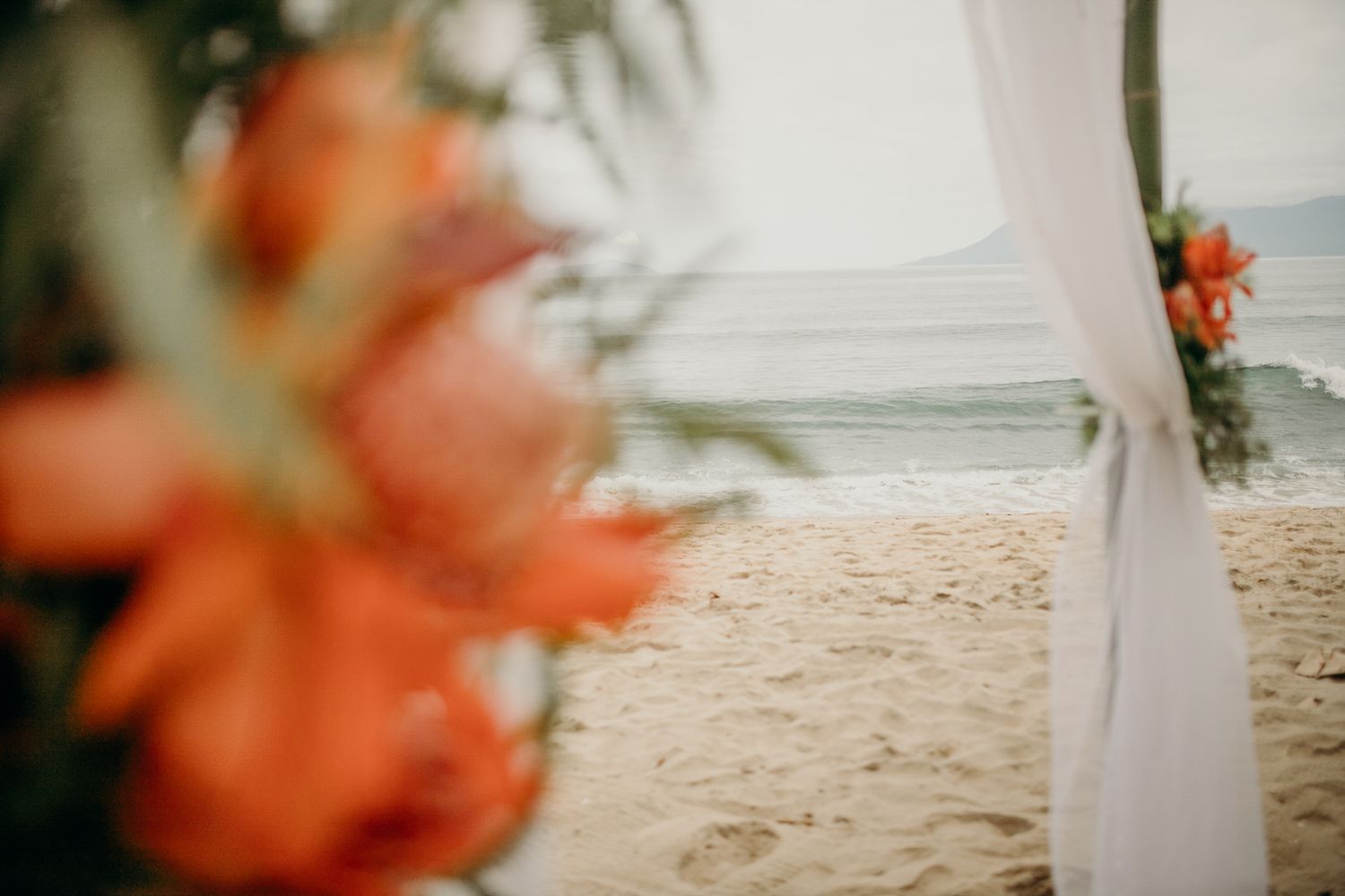 Beach wedding setup with white fabric draped posts and tropical floral arrangements.