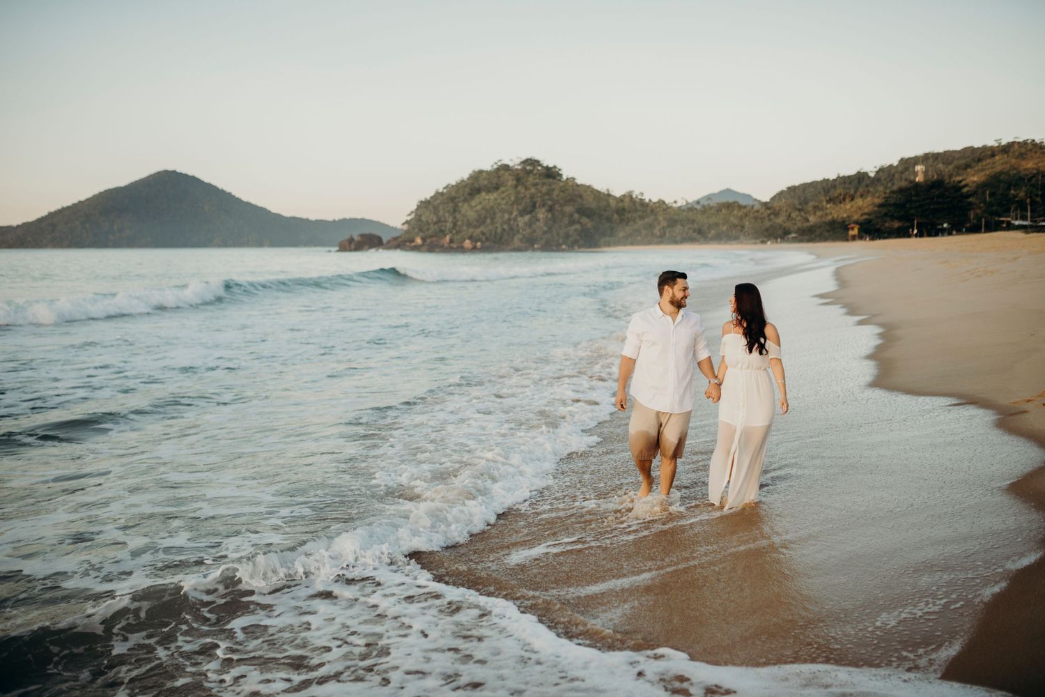 Couple walking along shoreline at sunset with mountains and waves in background.