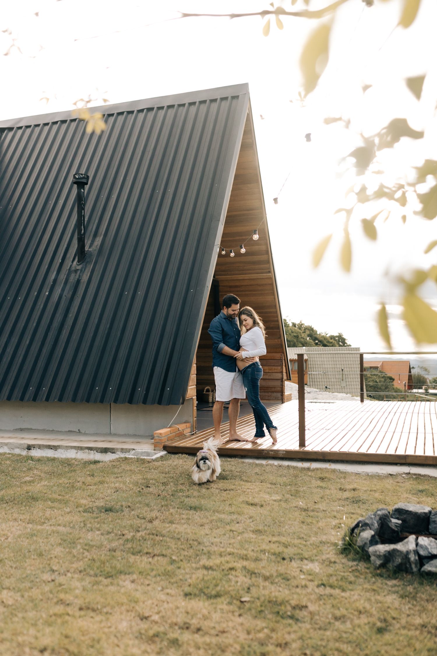 A couple poses together at an A-frame cabin with a dog nearby at sunset.