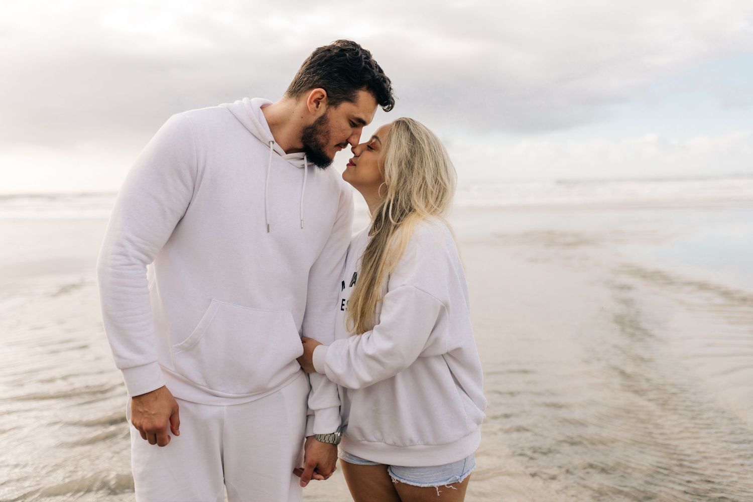 Intimate moment between couple wearing white outfits on a cloudy beach.
