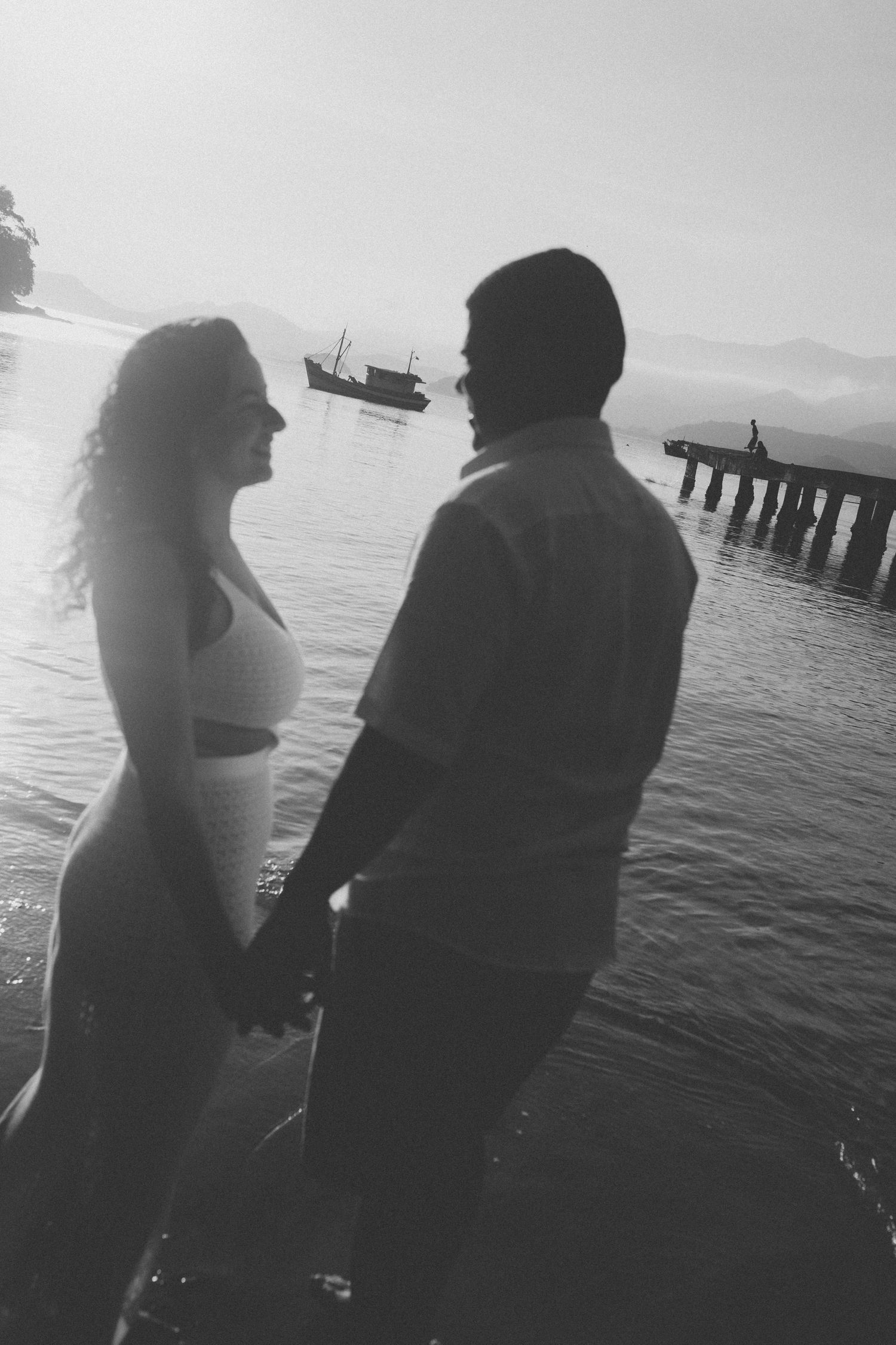 Couple in silhouette standing by waterfront during sunset with boats in background.