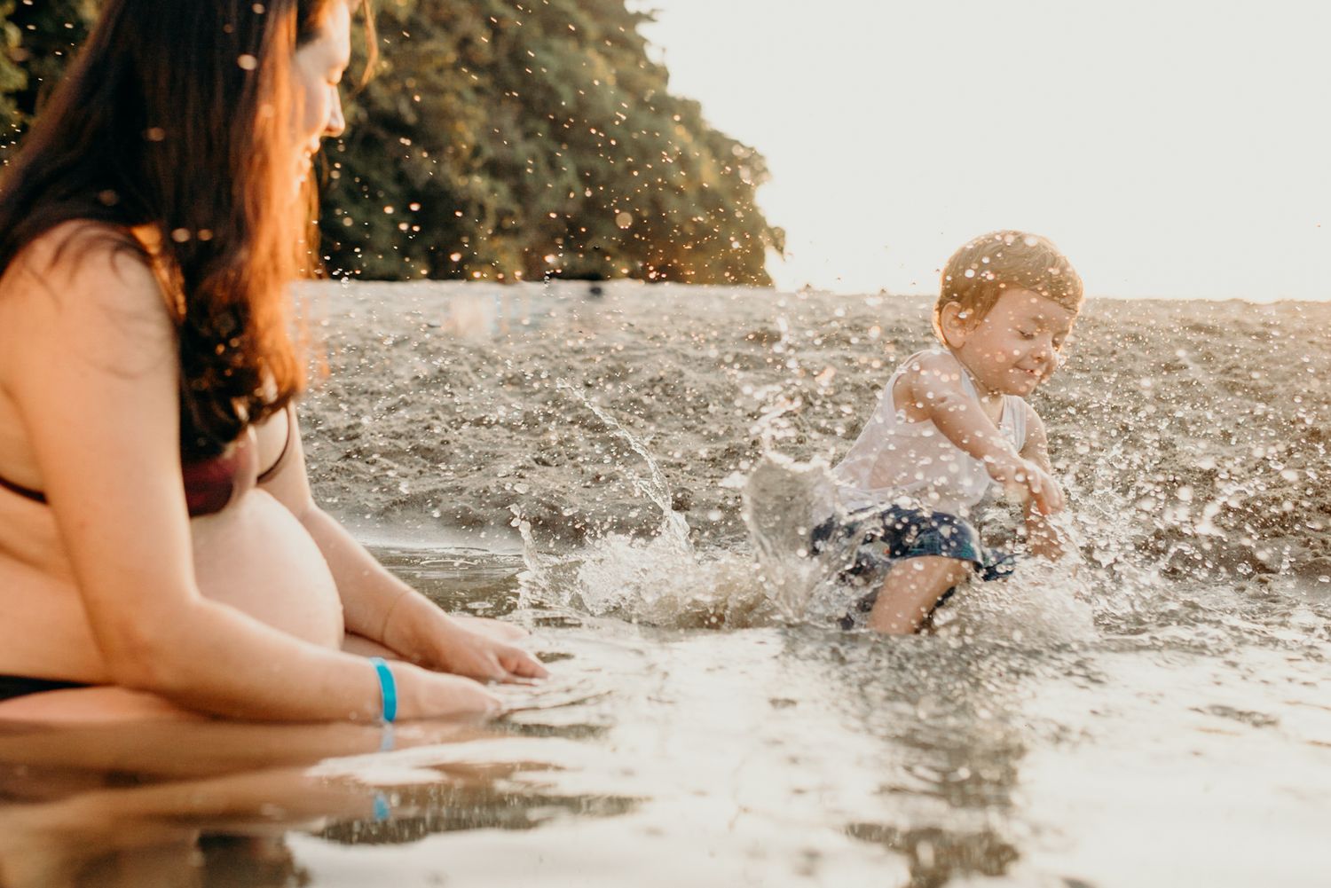 Child plays in beach water creating splashes while parent watches from nearby.