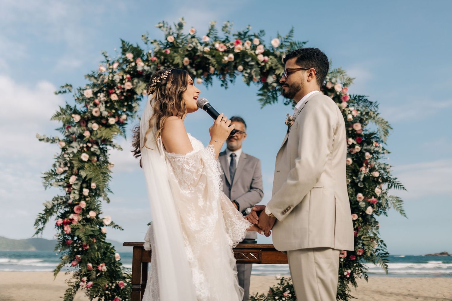 Intimate beach wedding ceremony with pink and white floral arch against blue sky.