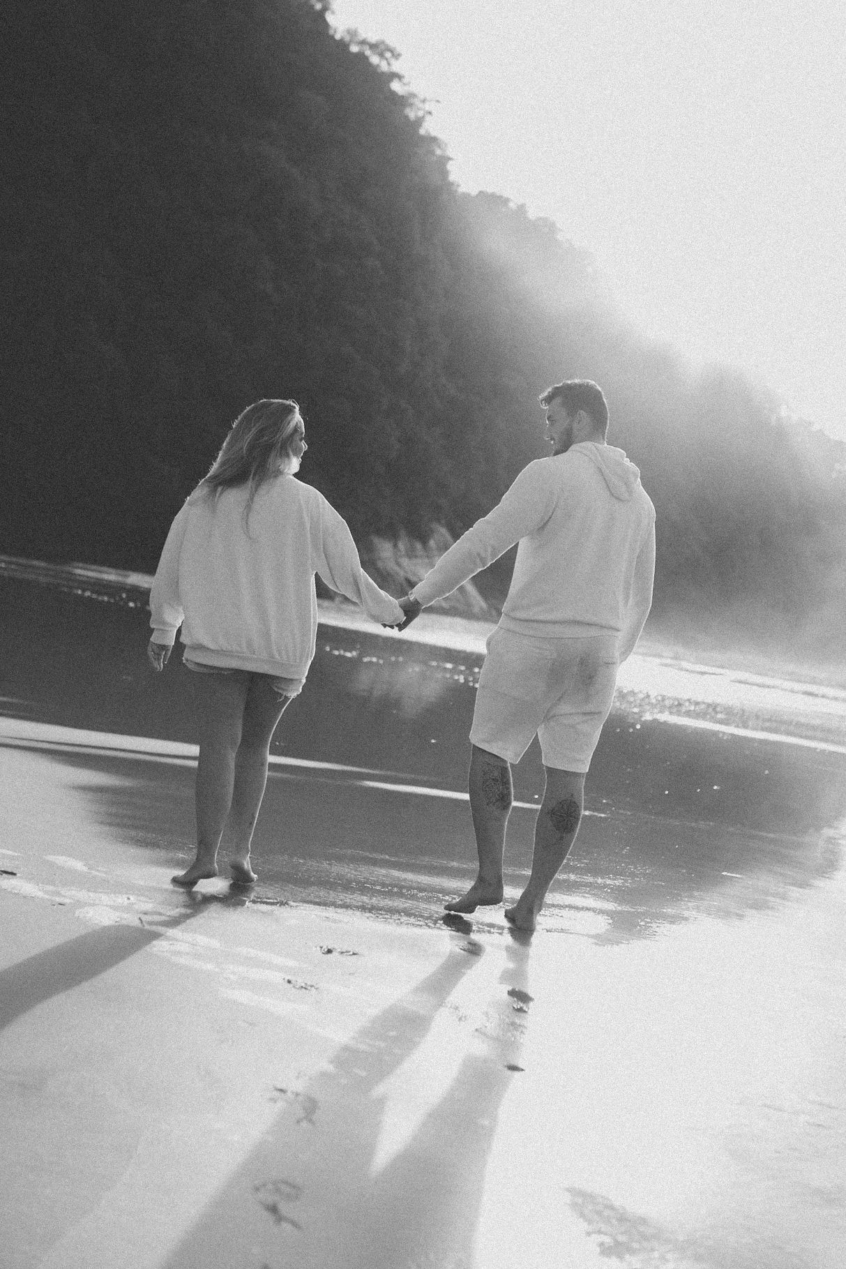Black and white photo of couple holding hands walking along misty beach.