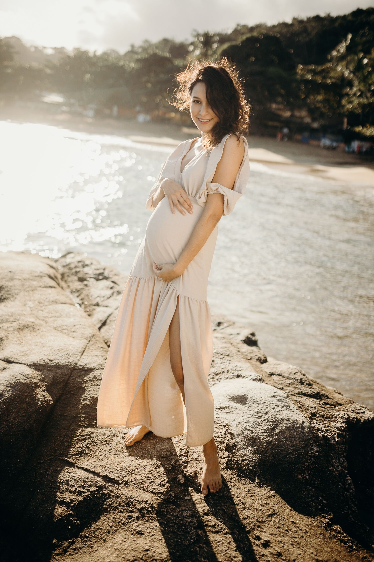 Backlit maternity photo on rocky beach showing expectant parent in flowing beige dress.