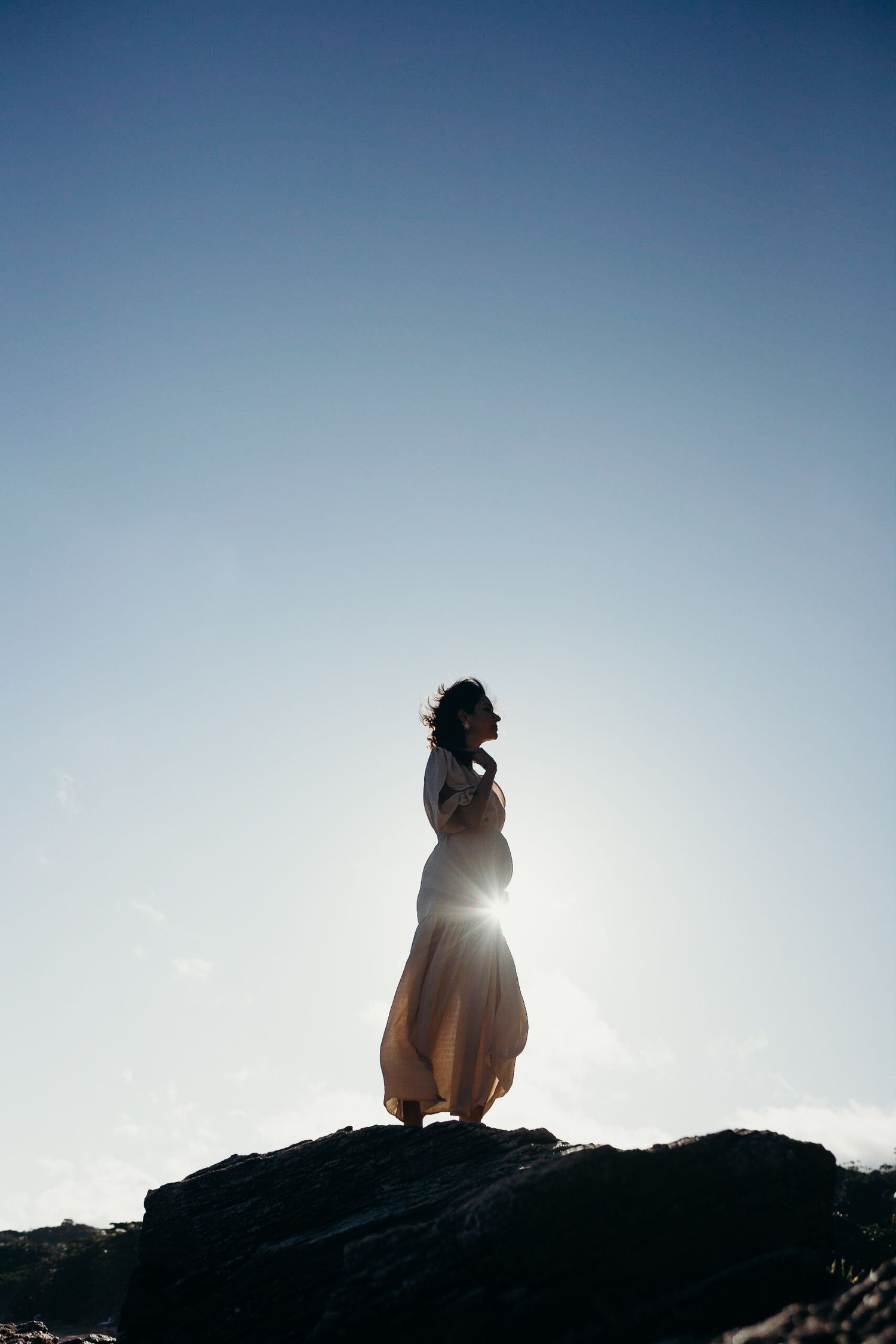 Silhouette of a figure in flowing dress standing on rocks against bright sky at sunset.