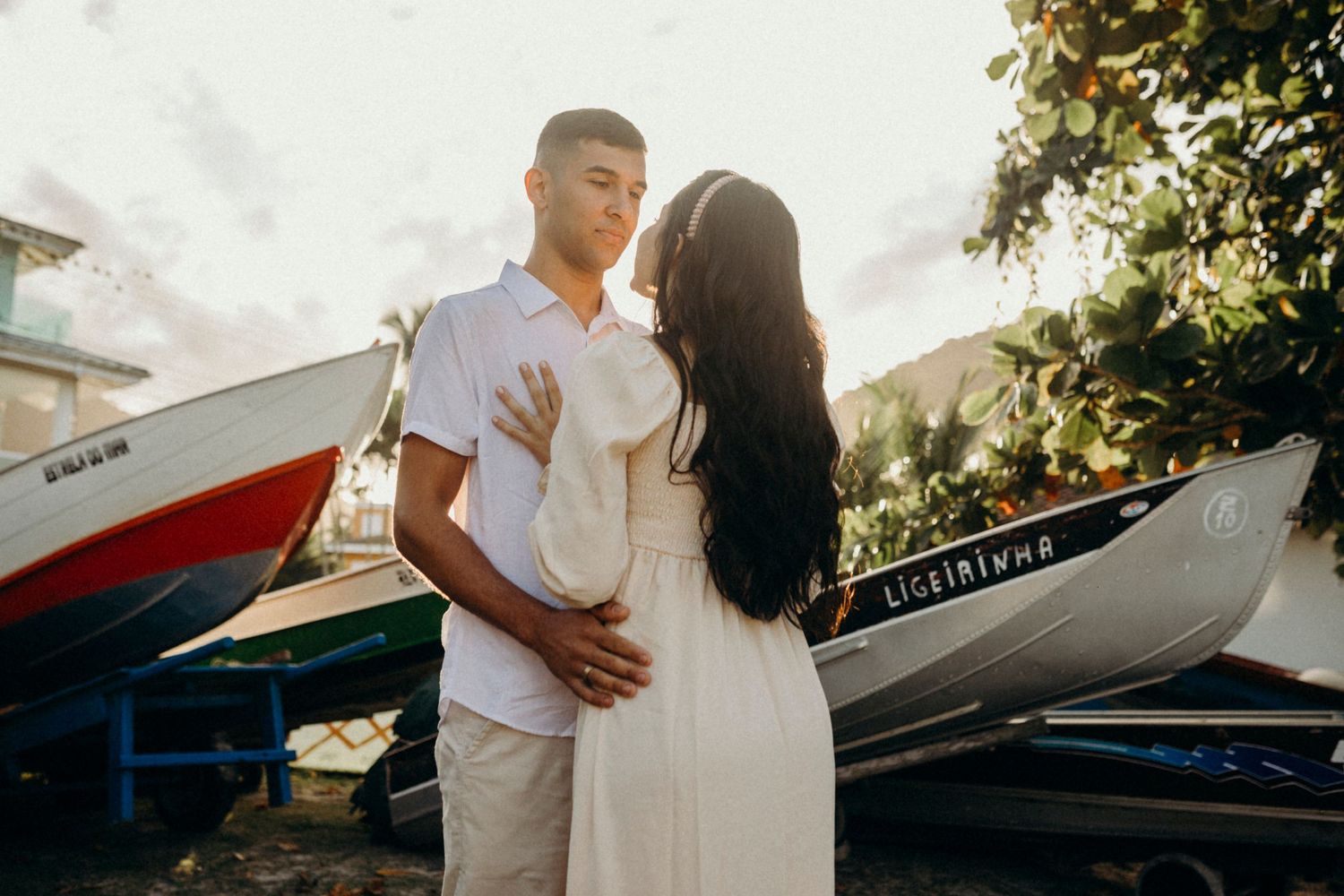 A couple in white outfits poses intimately near docked boats on a sunny day.