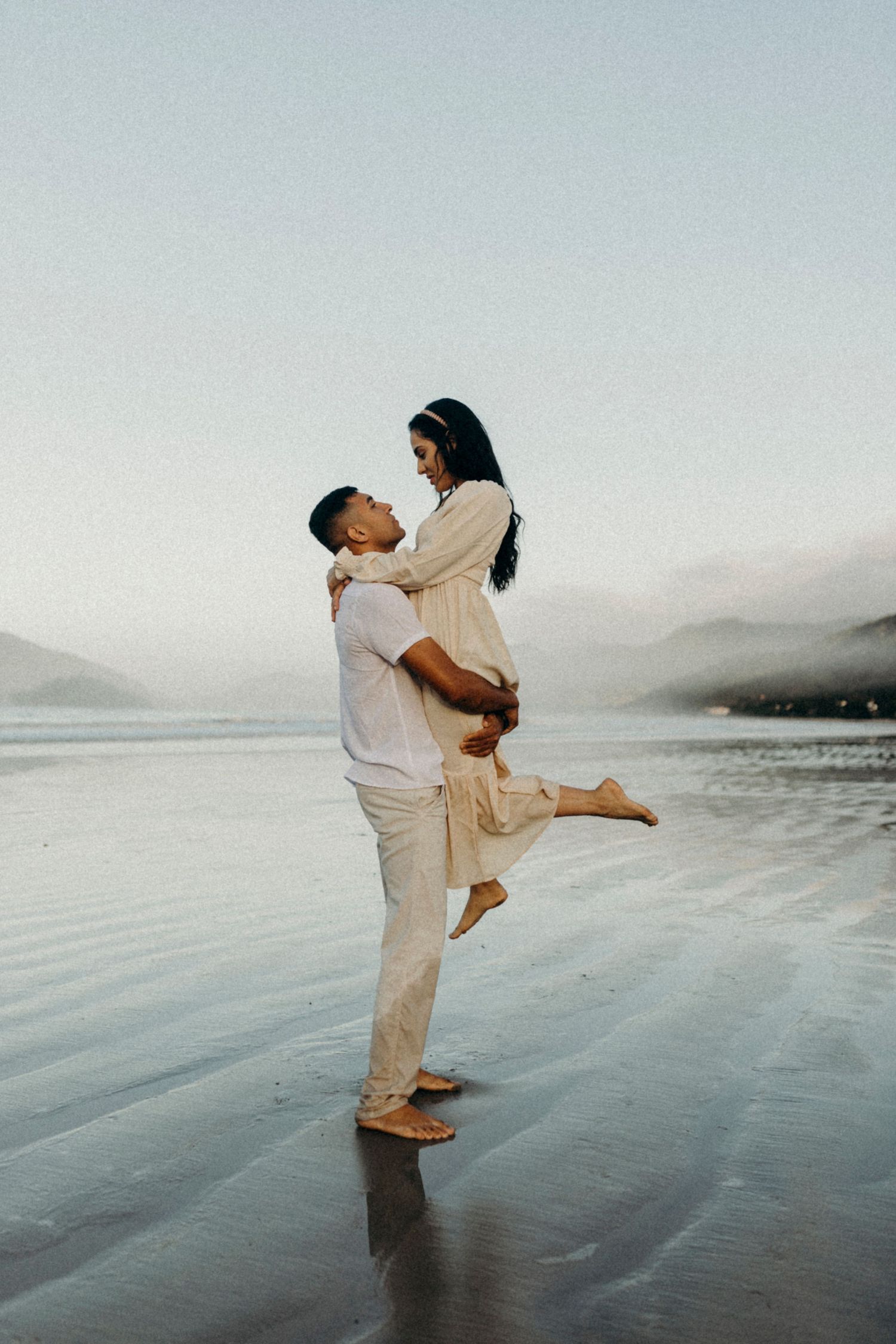 Couple on beach shares playful moment as one lifts the other in cream-colored outfits at sunset.