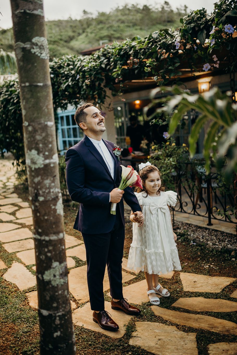 Formal event attendees stand on stone pathway under tropical trees during outdoor celebration.