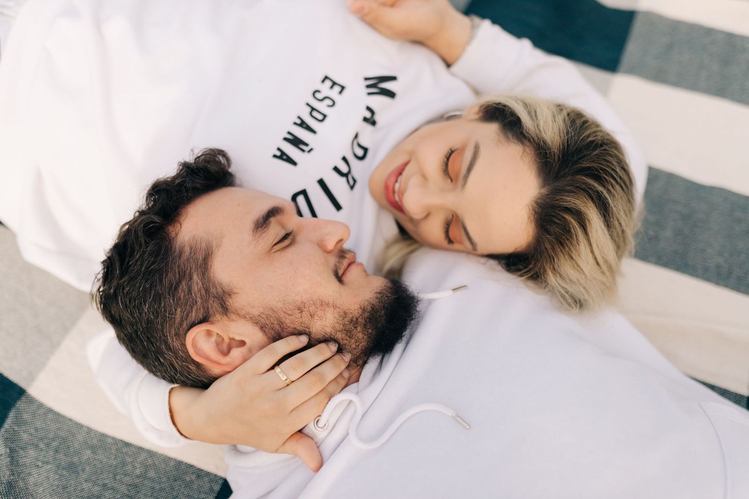 A couple lying on a striped blanket share an intimate moment in matching white outfits.
