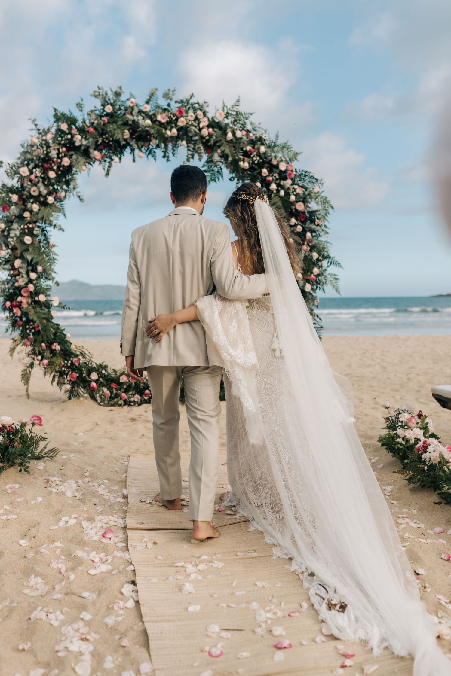 Back view of newlyweds standing under a circular floral arch at a beach ceremony.