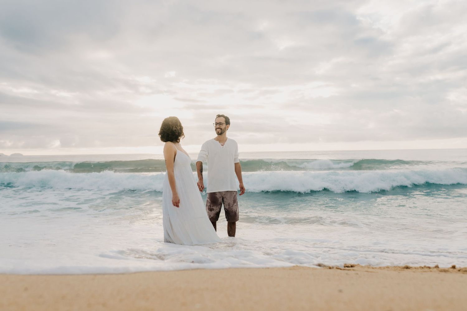 A couple in white attire stands on a beach with crashing waves behind them.