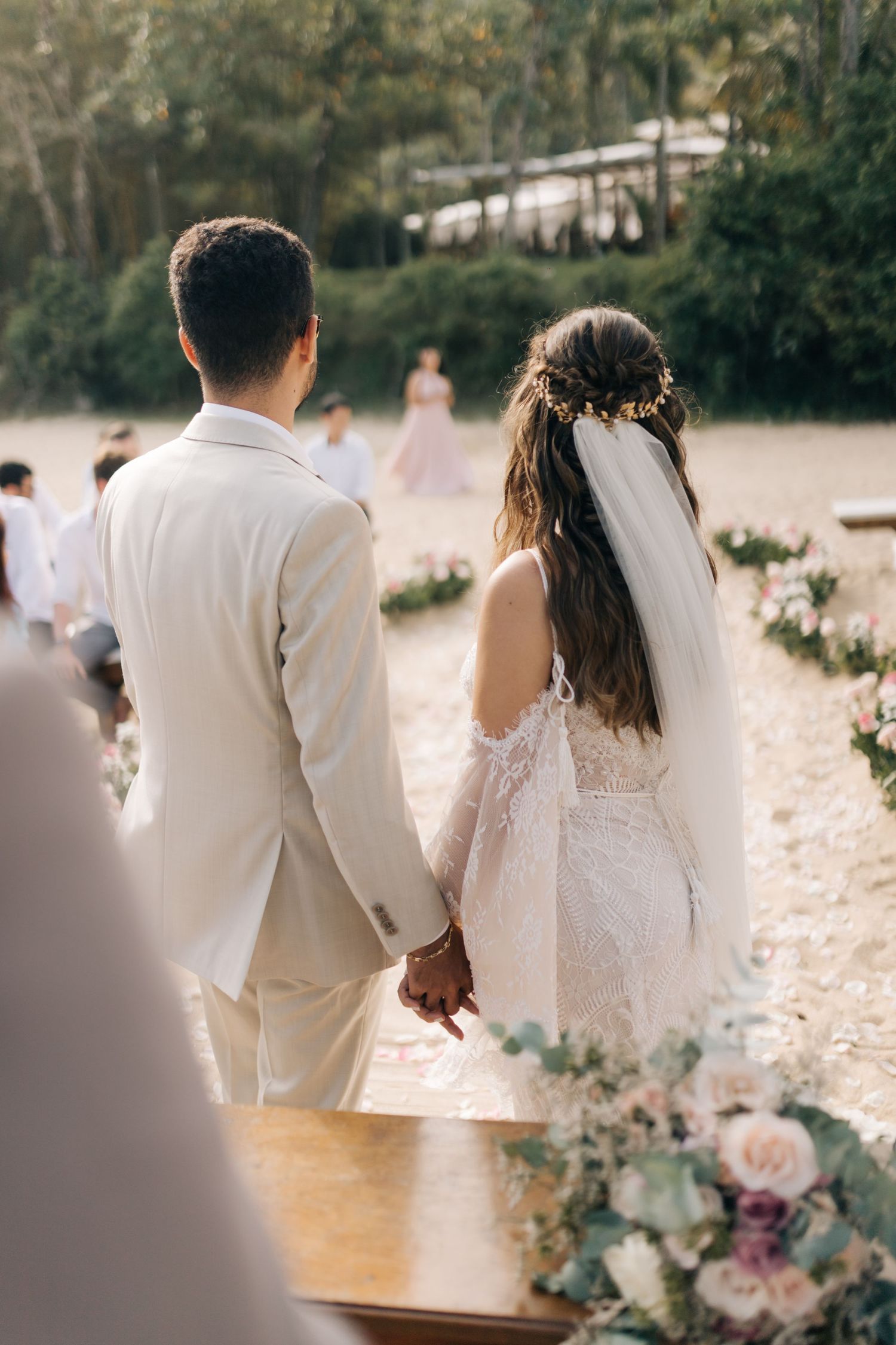 A couple holds hands during their beach wedding ceremony with the bride wearing a lace dress and veil.