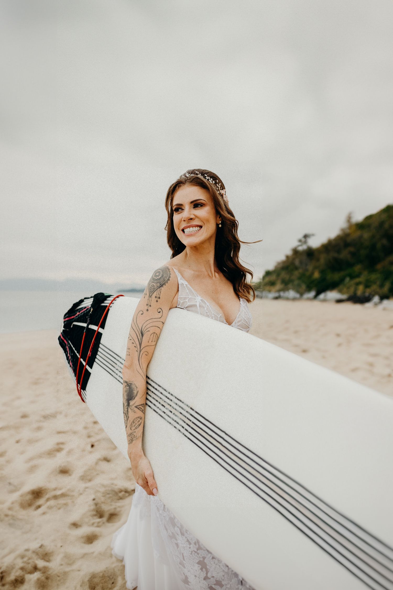 Woman in wedding dress holds a surfboard on sandy beach with ocean view.