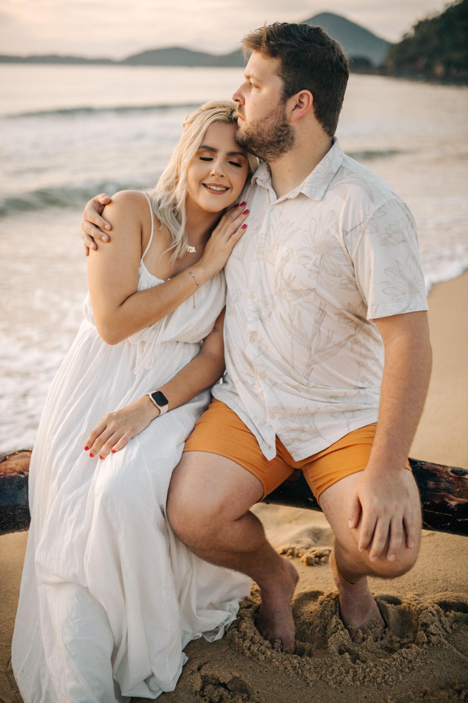 A couple embraces on a sandy beach at sunset with coastal mountains visible in the distance.