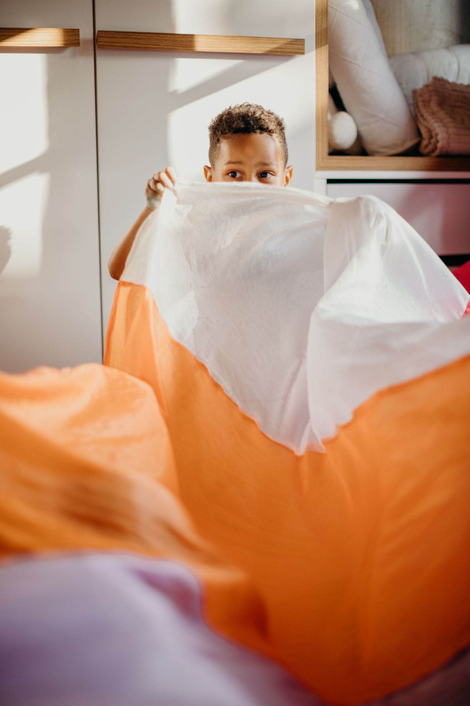 Child peeking playfully from behind white and orange fabric sheets.