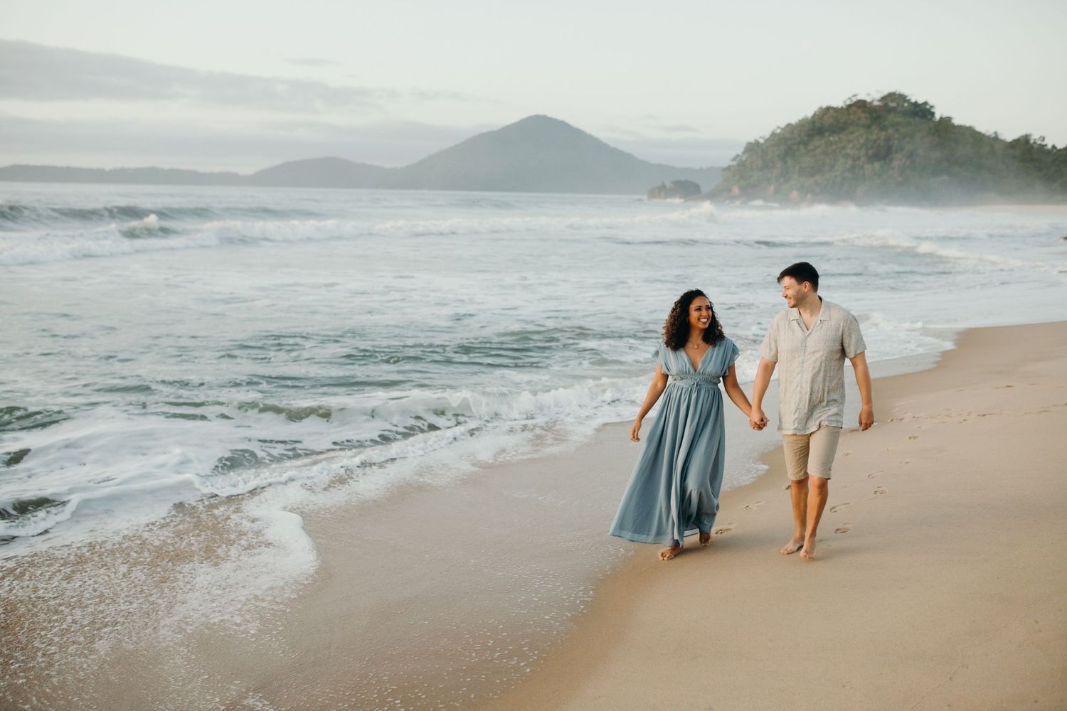 Couple walking hand in hand along scenic beach with mountains and ocean waves.
