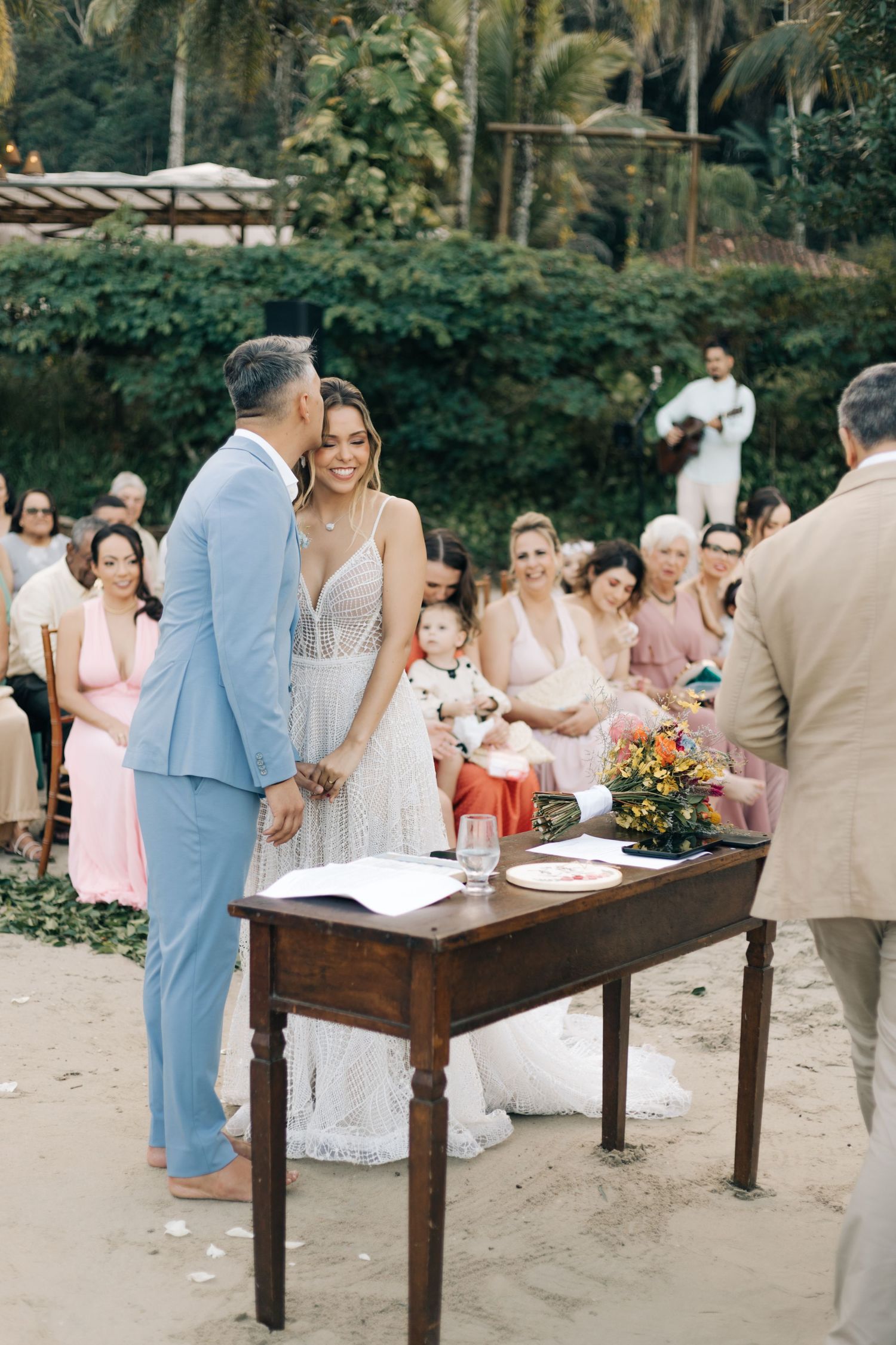 Outdoor wedding ceremony with guests seated in pink attire and a rustic wooden table setup.