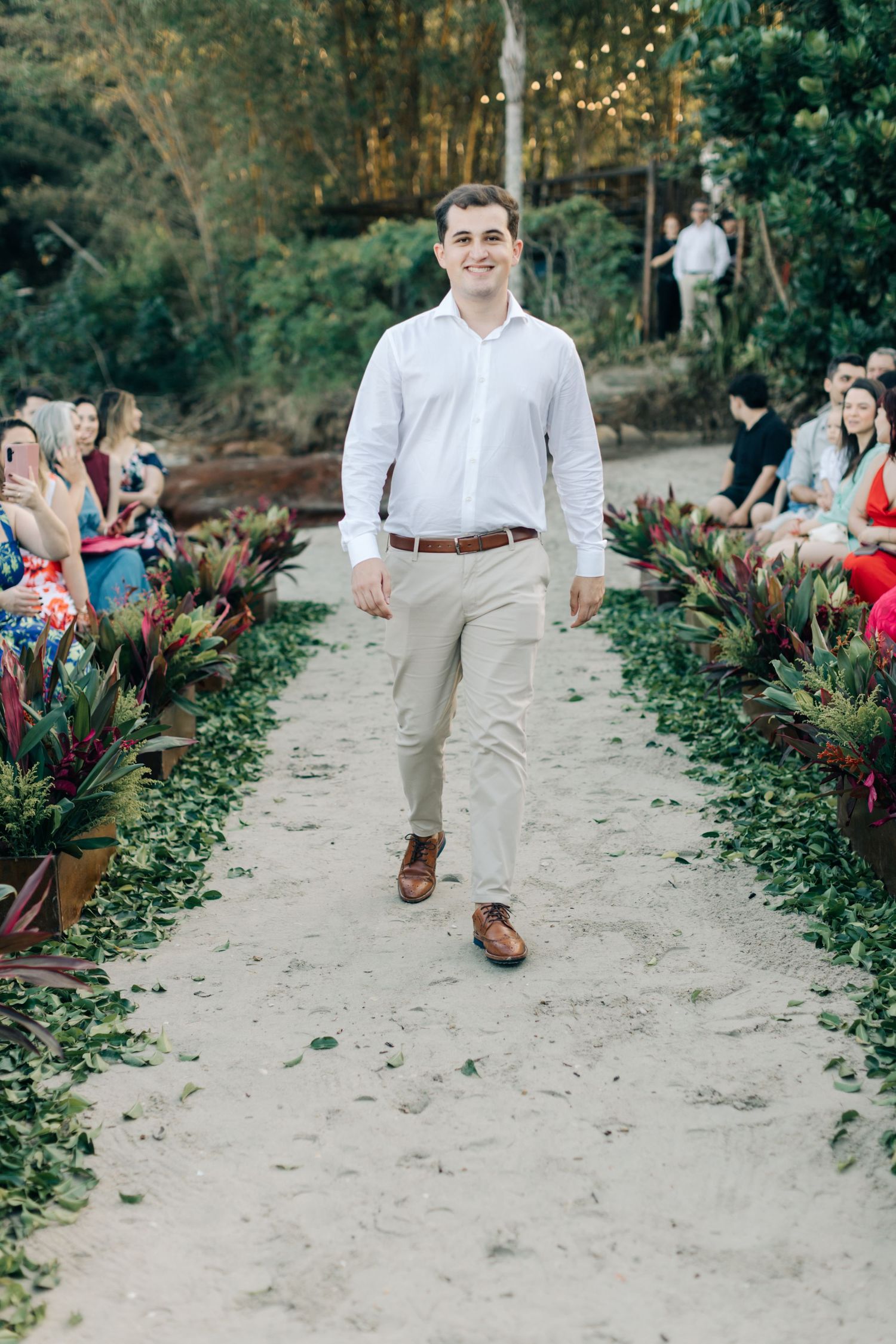 Wedding party member in crisp white shirt and khakis walks sandy aisle at outdoor ceremony.