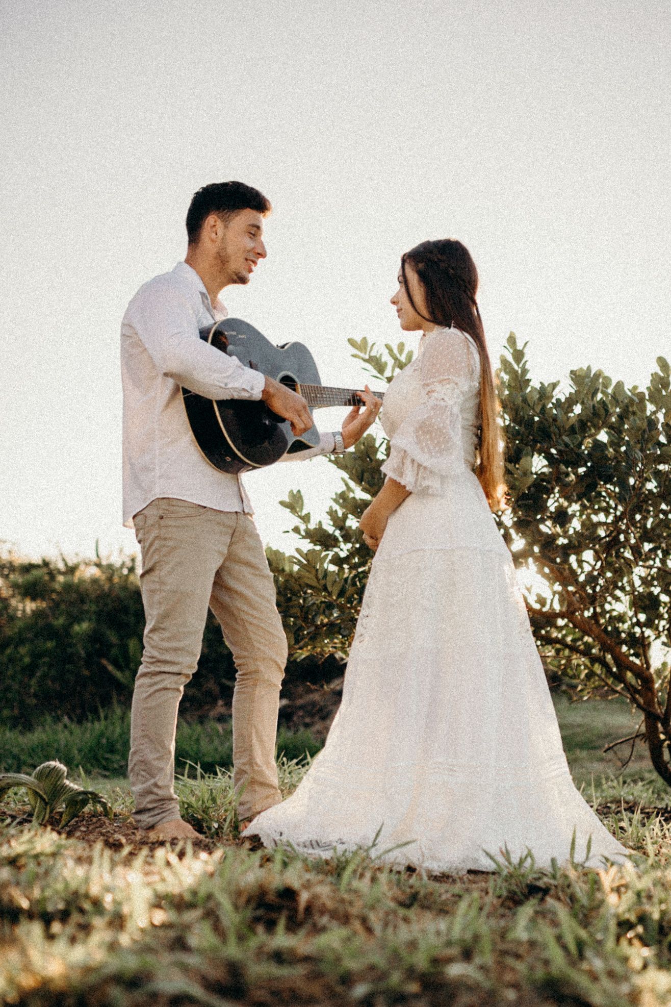 Couple in wedding attire share a joyful moment with acoustic guitar outdoors.