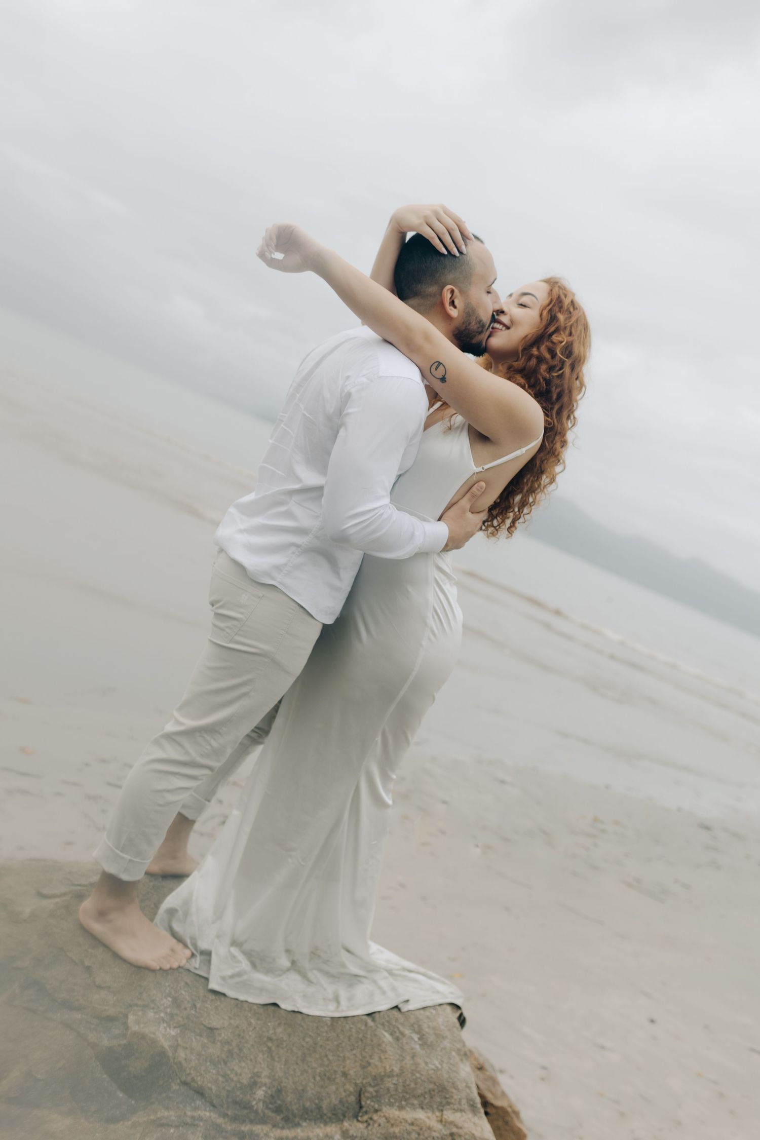 A romantic beach photoshoot captures a couple in white attire sharing passionate moments.
