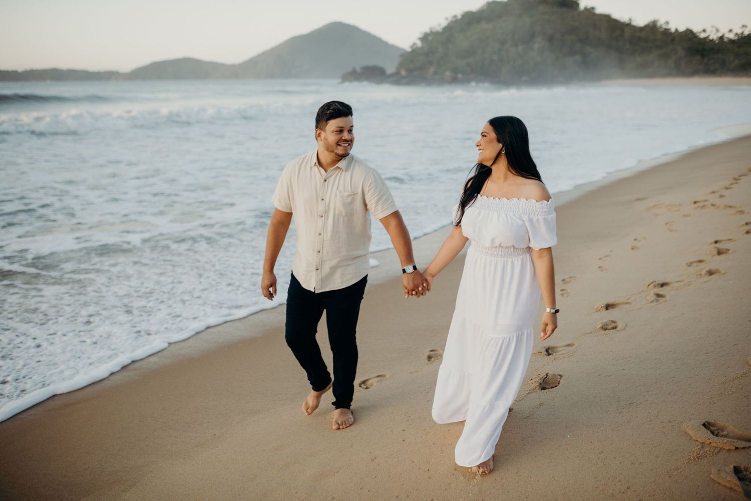 Couple strolling together on a sandy beach with waves and mountains in view.