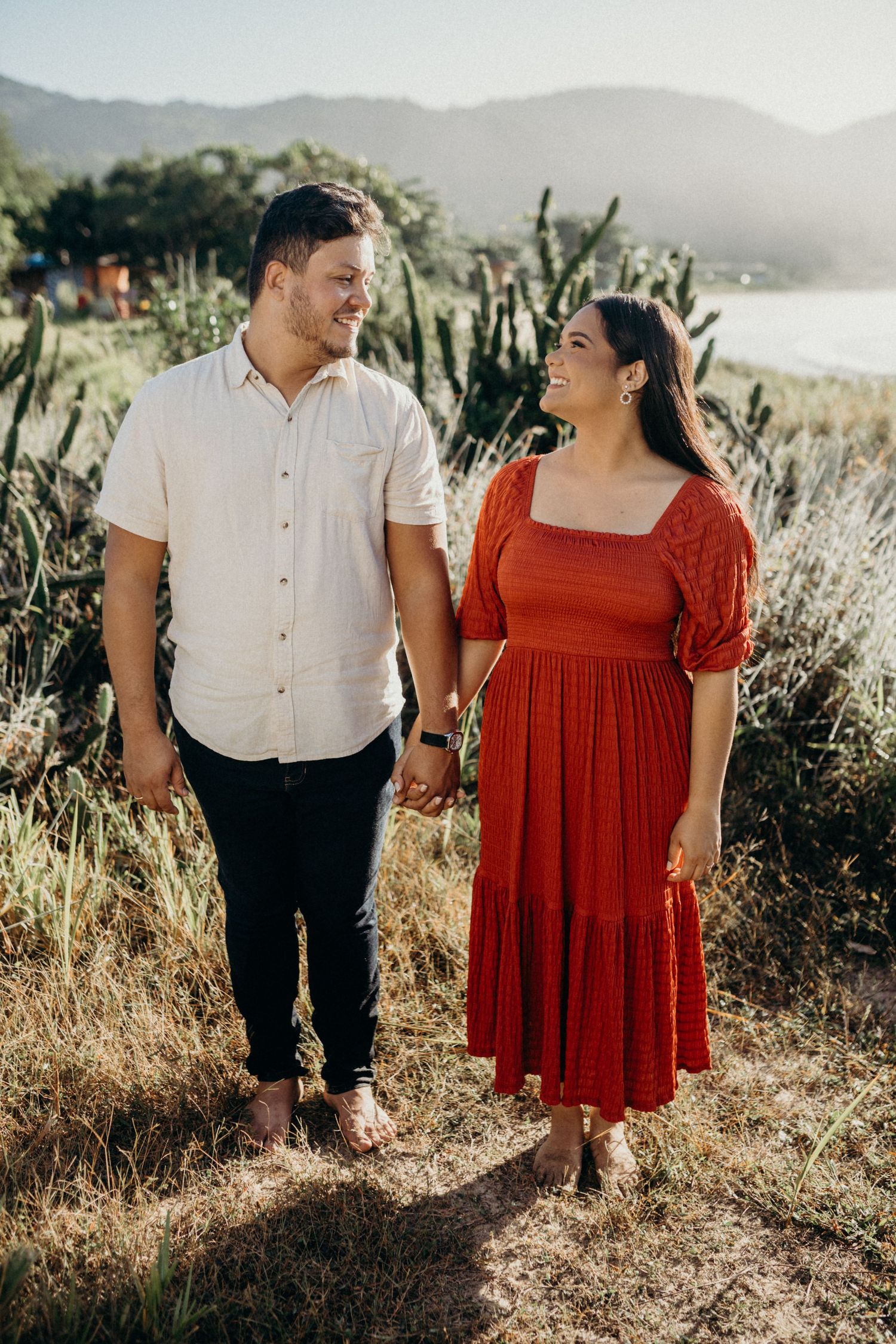 A couple holding hands stands on a hillside with cacti and mountains in the background during golden hour.