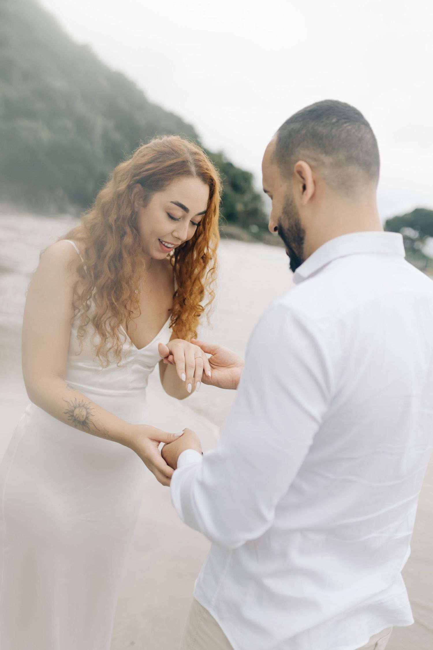 A couple shares an intimate moment on a misty beach as they look at a ring during their engagement.