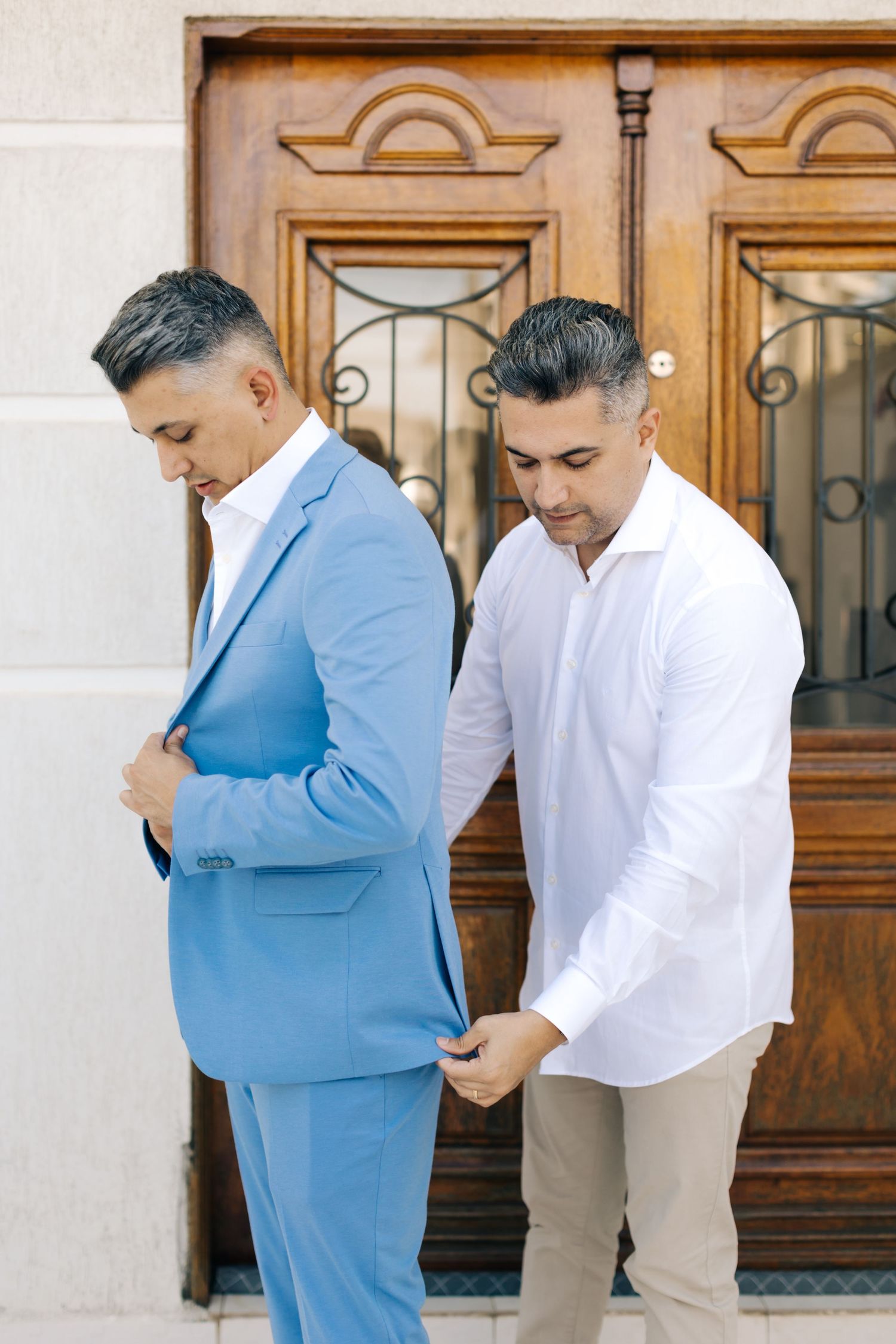 Two men in formal attire adjusting suit outside ornate wooden doors.
