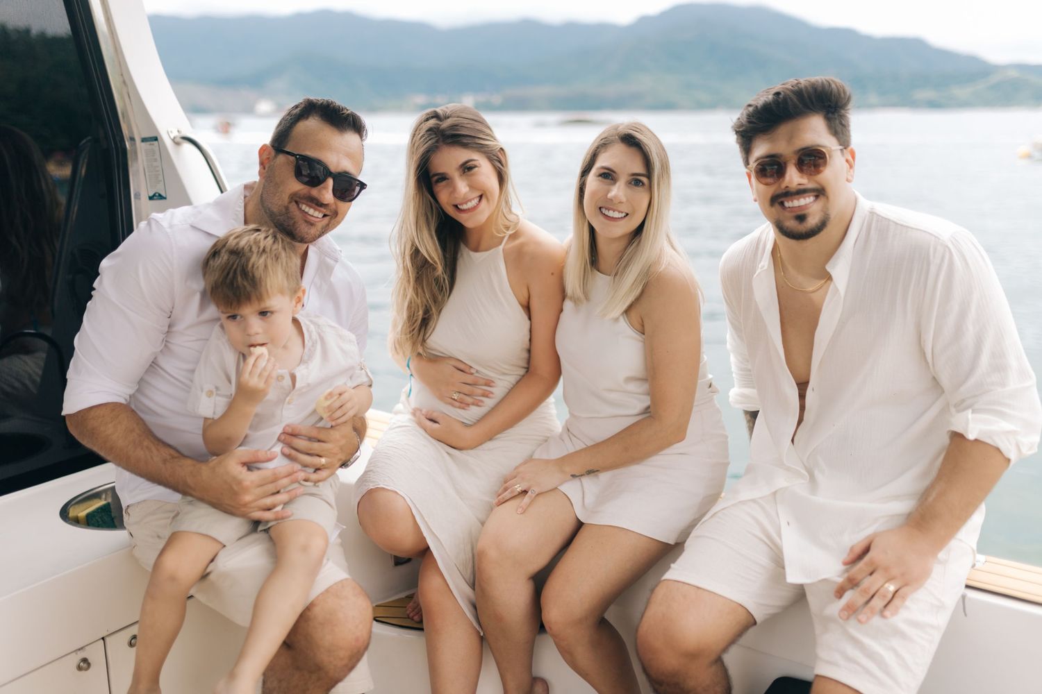 Family wearing white outfits poses together on boat deck with scenic mountain backdrop.