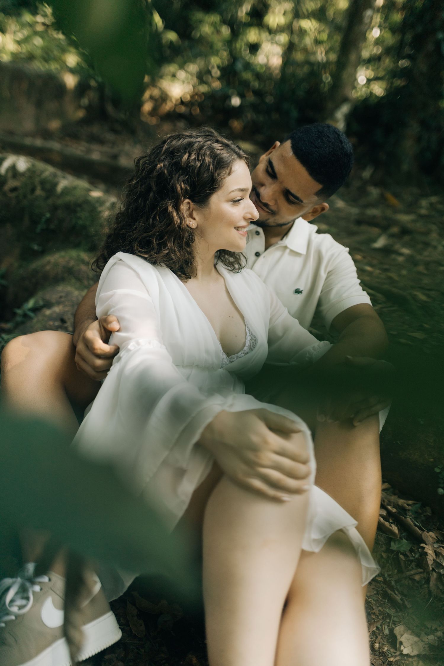 Romantic couple in white outfits share intimate moments in a wooded setting with natural light filtering through trees.