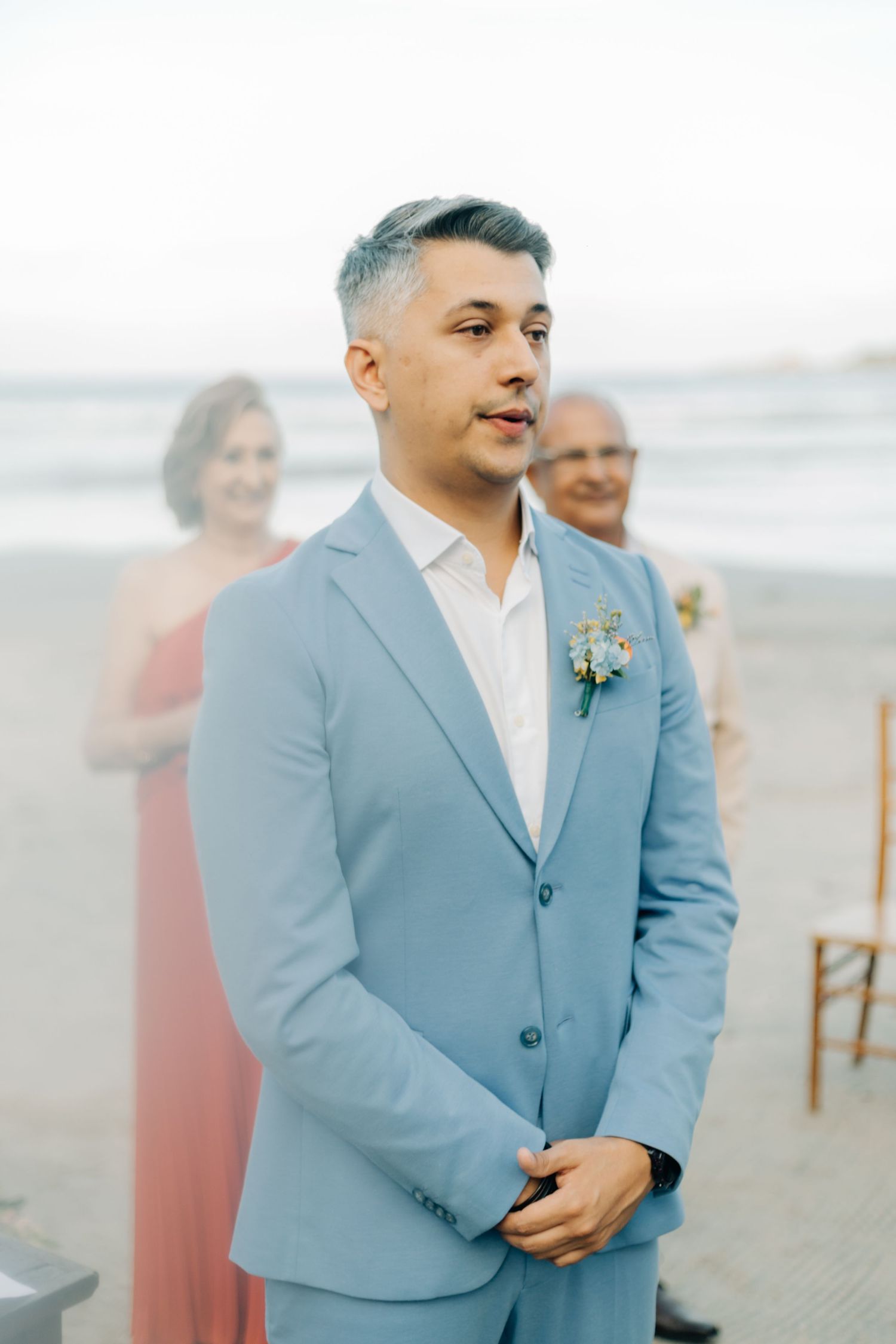 A groom in a light blue suit stands on a beach during a seaside wedding ceremony.