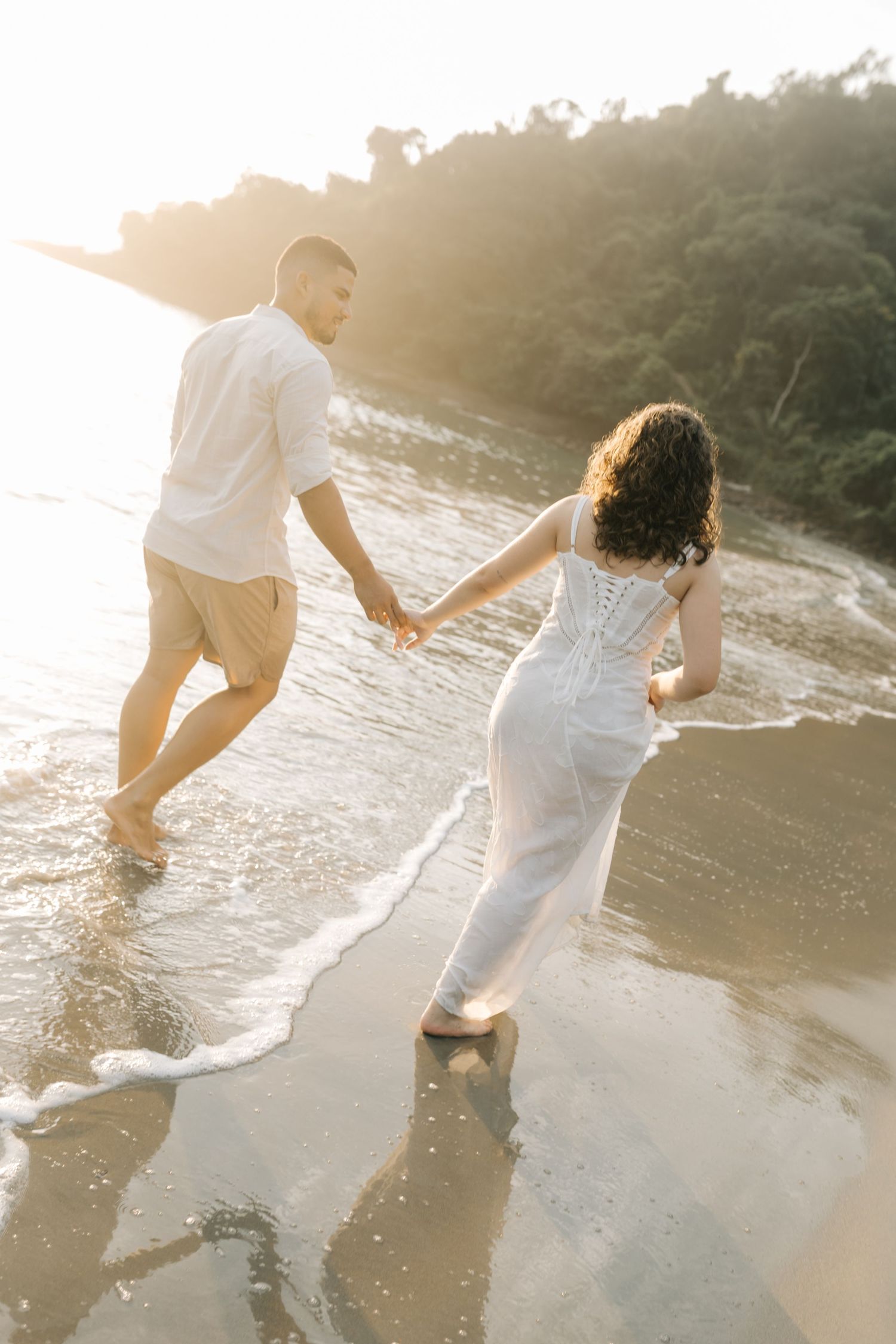 Couple walking together on wet sand as waves crash on a beach at sunset.