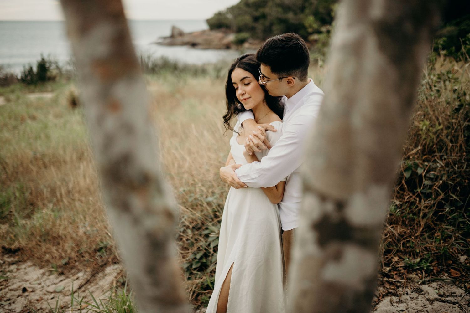 A couple sharing a romantic moment framed by tree trunks with ocean views in the background.
