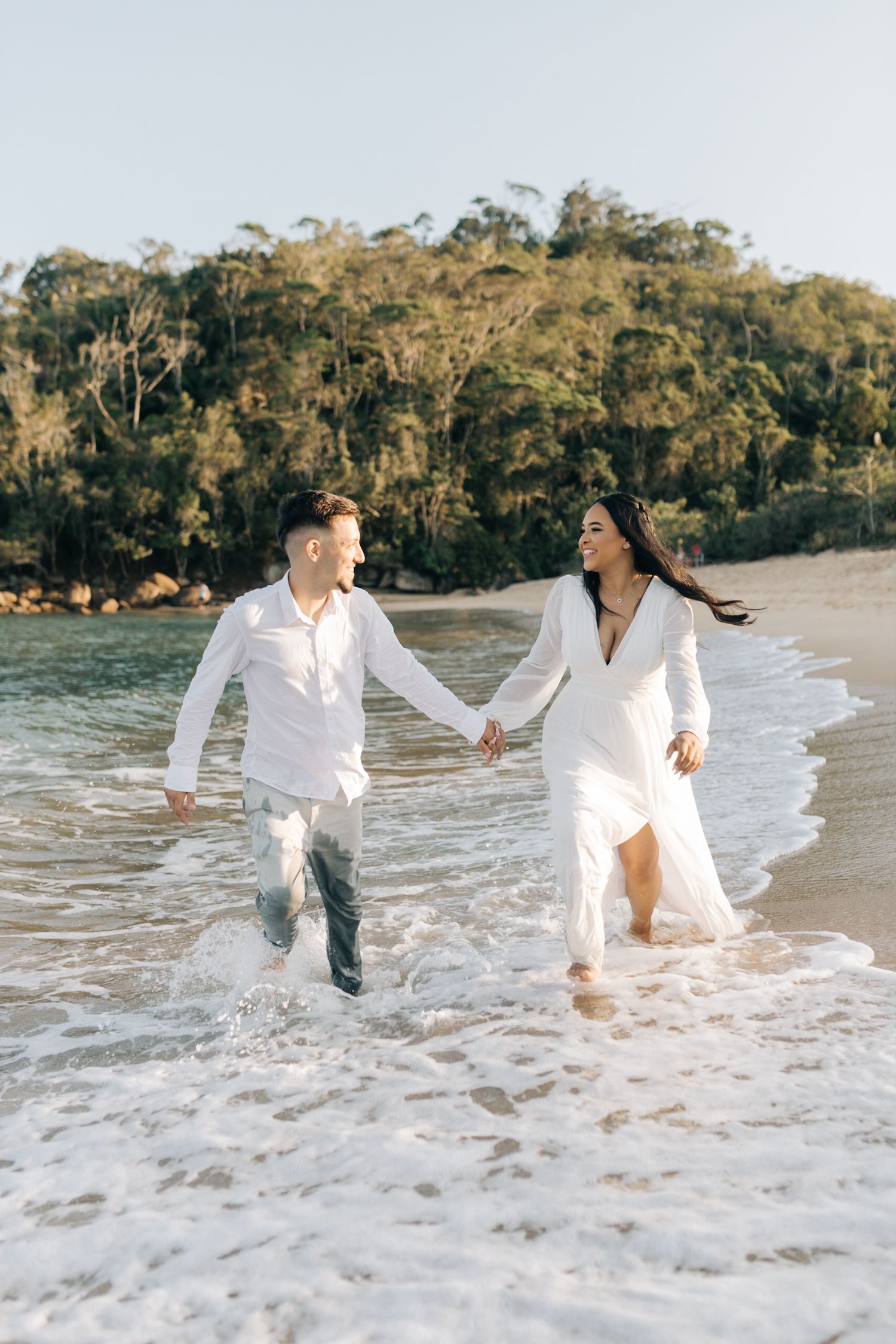Couple runs and plays in ocean waves wearing matching white outfits at scenic beach.